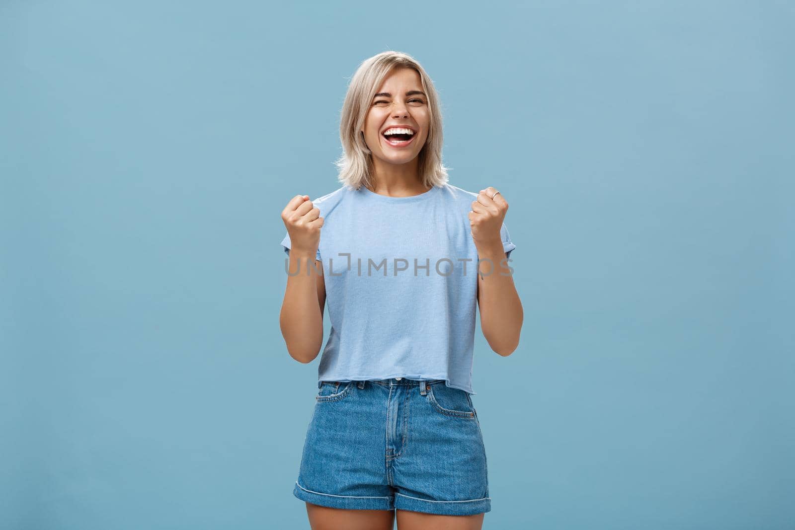 Lucky happy girl triumphing and laughing from happiness winning first place clenching fists in success gesture smiling broadly celebrating victory with enthusiasm and excitement over blue background. Emotions concept