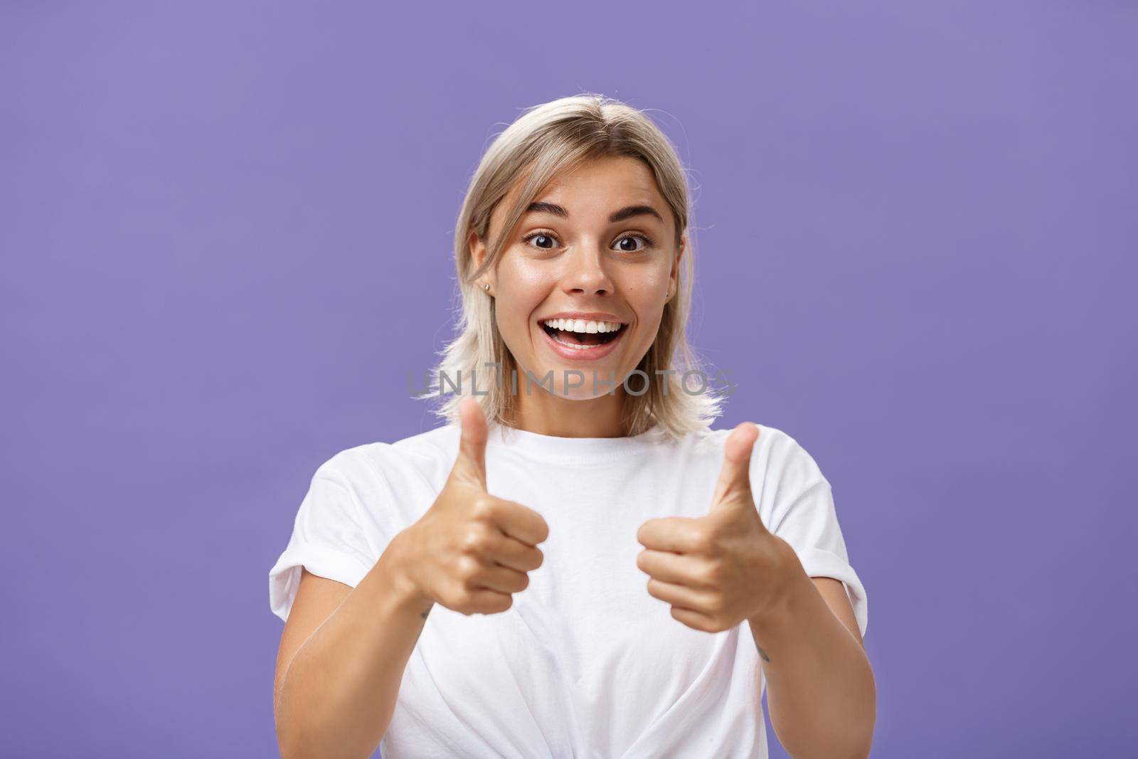 Waist-up shot of optimistic and supportive good-looking caucasian female in white t-shirt cheering and smiling broadly hearing awesome idea approving it with thumbs up gesture over purple wall by Benzoix