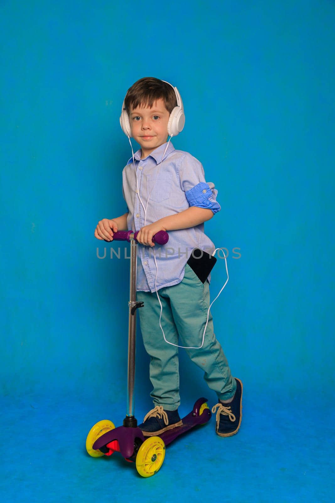 A boy on a scooter with headphones on a blue background . An article about children's scooters. An article about the choice of a scooter for a child. An article about the safety of the child. A happy child. Blue background and a boy. Copy Space