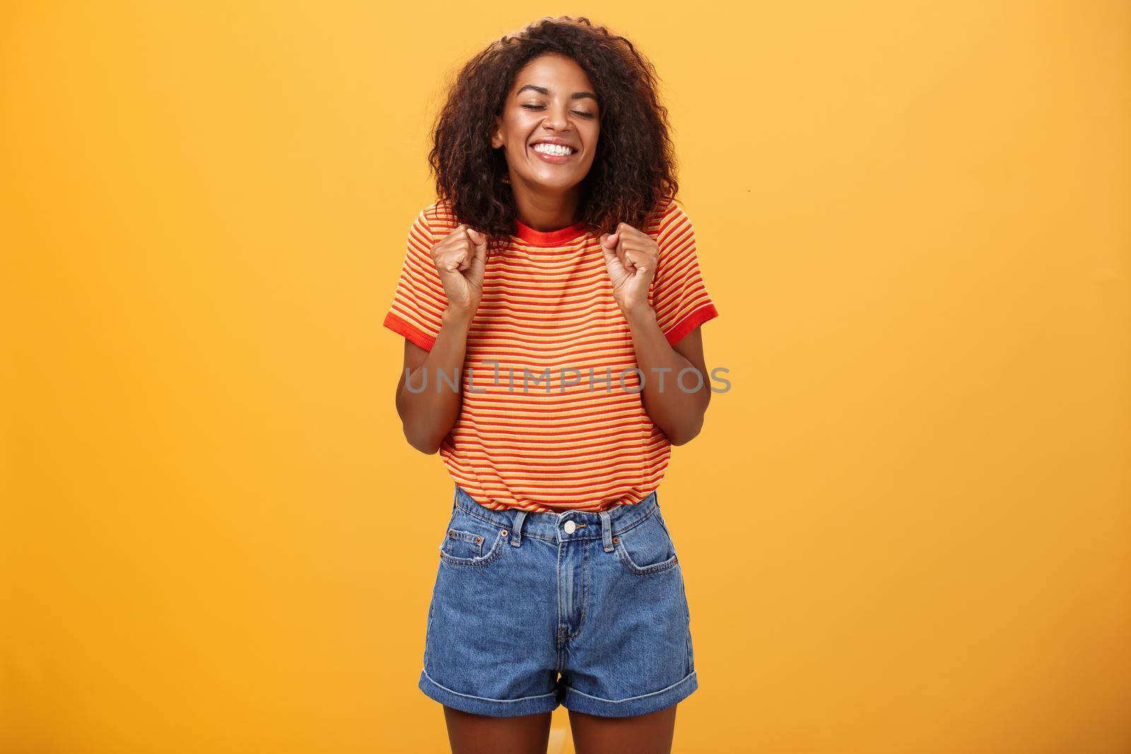 Girl eager to see lover coming from trip standing excited and thrilled with happy broad smile clenching fists grinning with closed eyes feeling joyful and amused over orange background. Lifestyle.