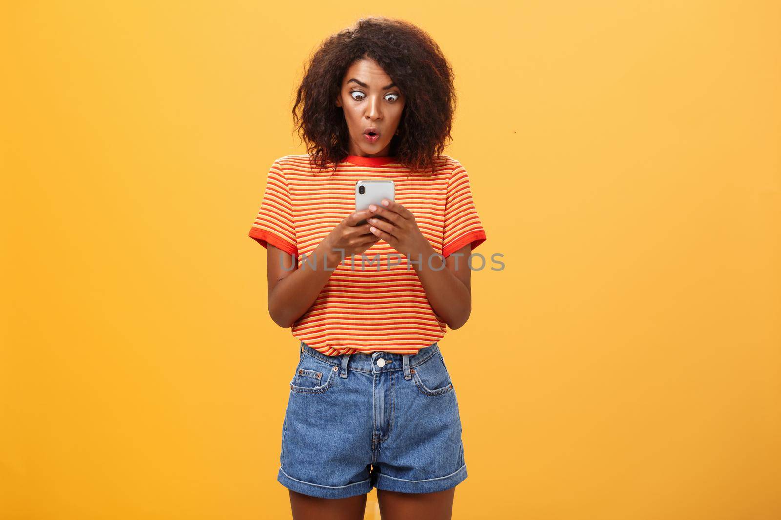 Indoor shot of shocked stunned african american young woman with afro hairstyle staring surprised and excited at smartphone screen holding cellphone reading amazing message over orange wall by Benzoix