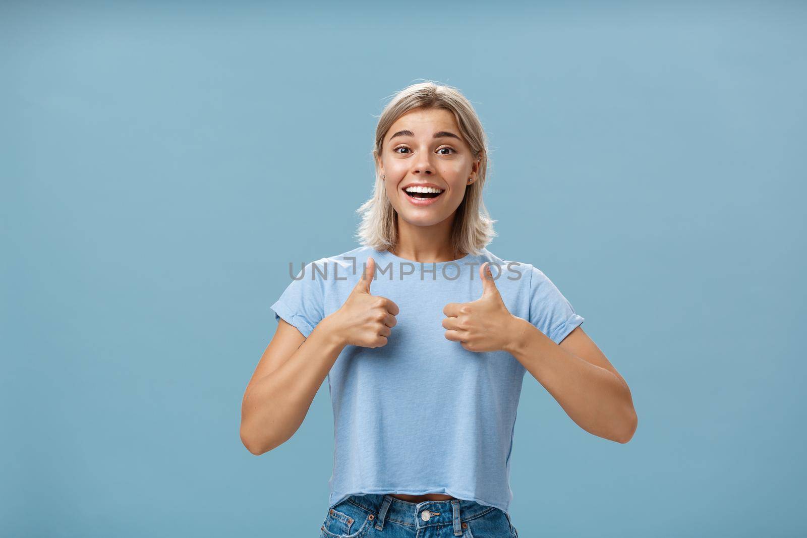 You did great proud of you. Portrait of satisfied and fascinated attractive happy girlfriend in casual t-shirt showing thumbs up and smiling broadly being supportive and cheerful over blue wall by Benzoix