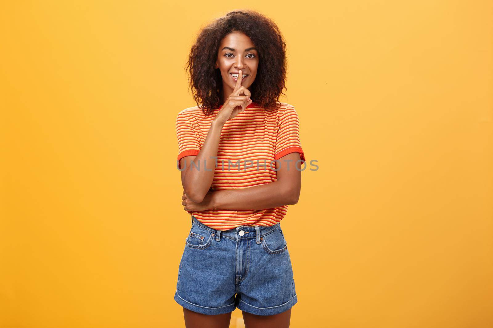 Keep secret inside I trust you. Feminine and sensual good-looking stylish dark-skinned woman with curly hair in trendy outfit shushing at camera with delighted mysterious look hiding something. Body language concept