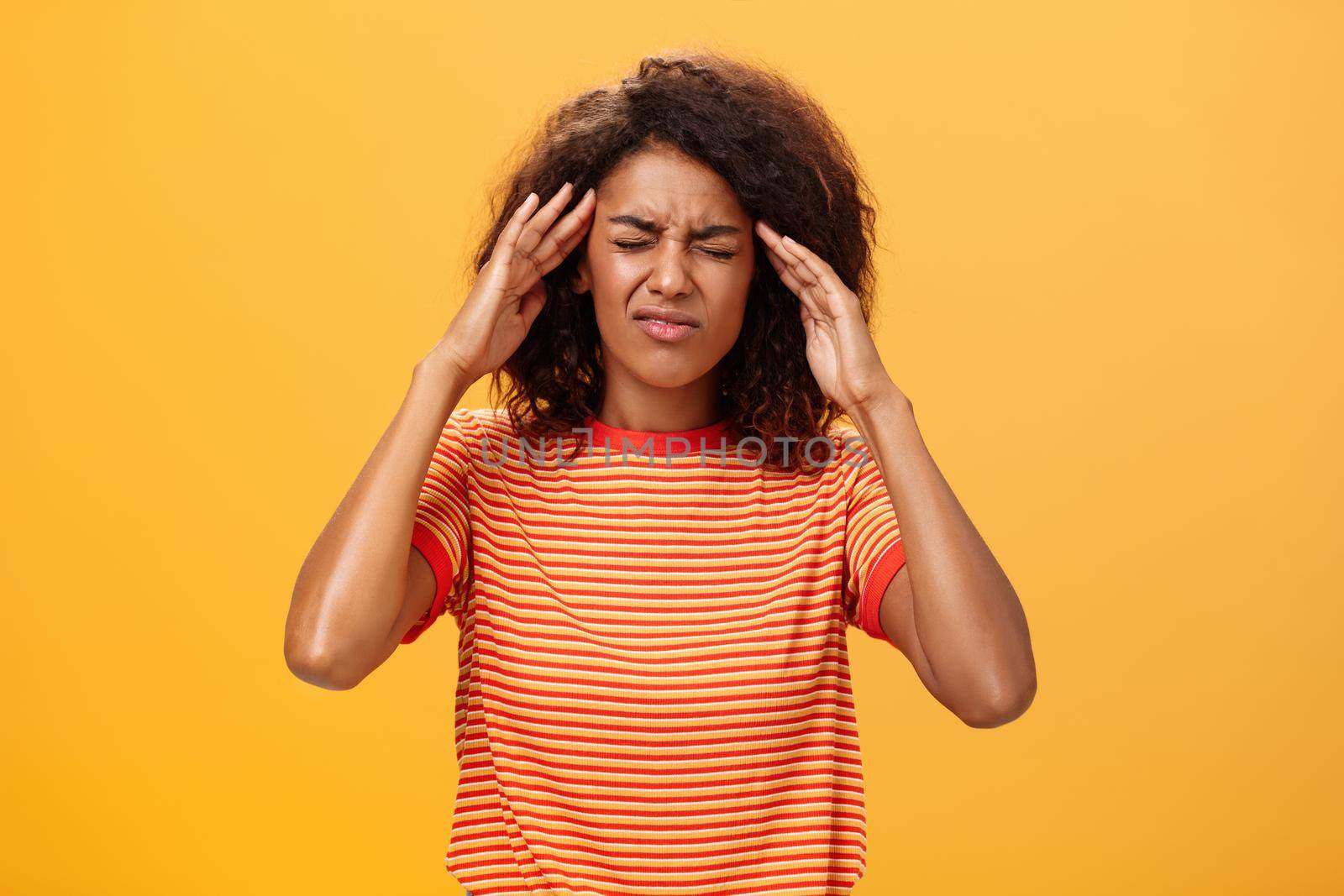 Woman cannot recall important information looking intense trying concetrate under pressure feeling tensed touching temples closing eyes while thinking hard posing against orange background.