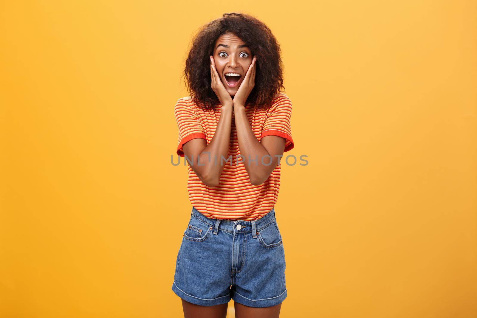 Impressed talkative african american woman finding out hot details of rumor being excited, thrilled leaning face on palms, smiling broadly with amused expression listening carefully over orange wall.