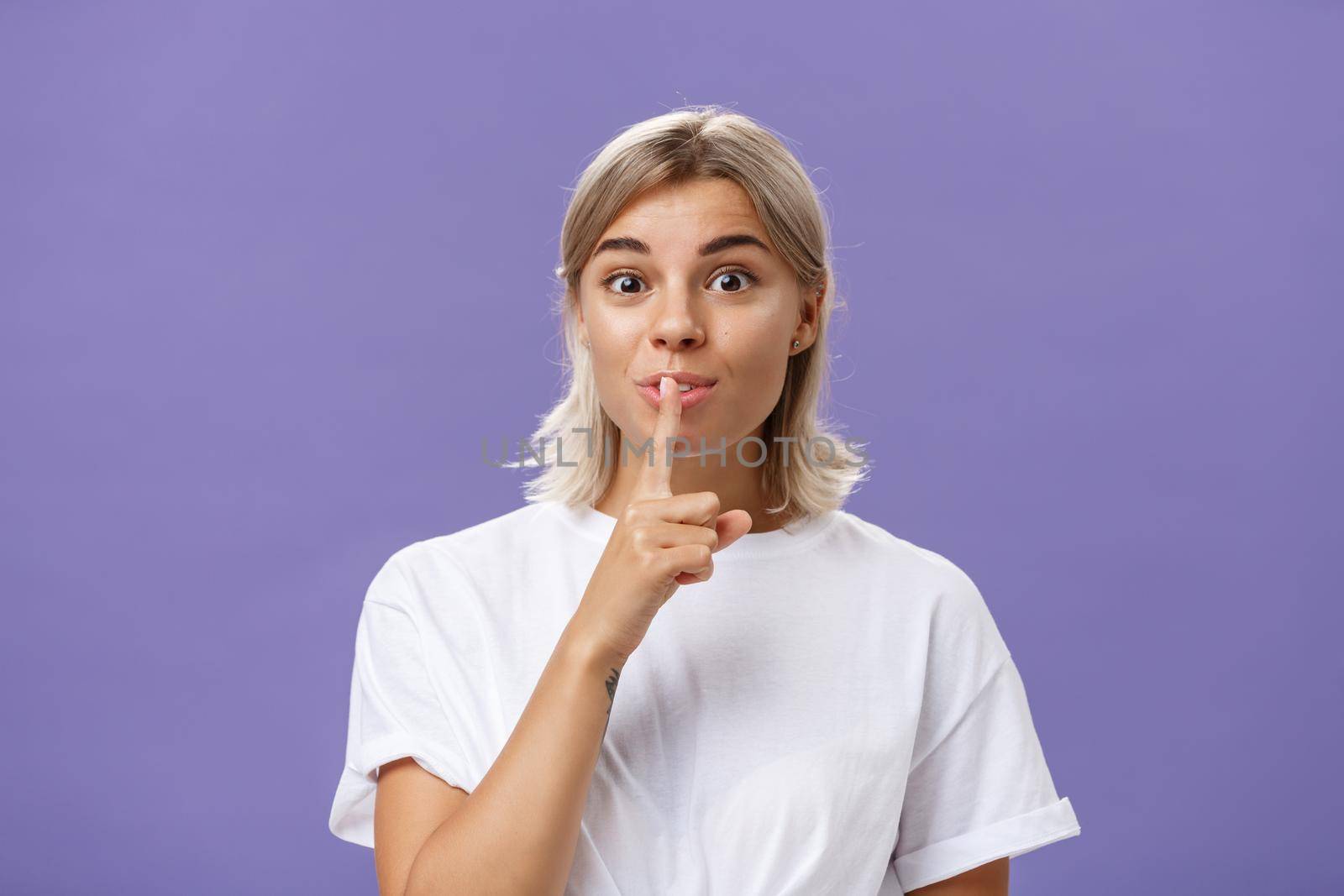 Lifestyle. Charming creative and stylish beautiful tanned girl with fair hair shushing at camera with delighted thrilled look making up great idea wanting hide it and keep secret posing over purple wall.