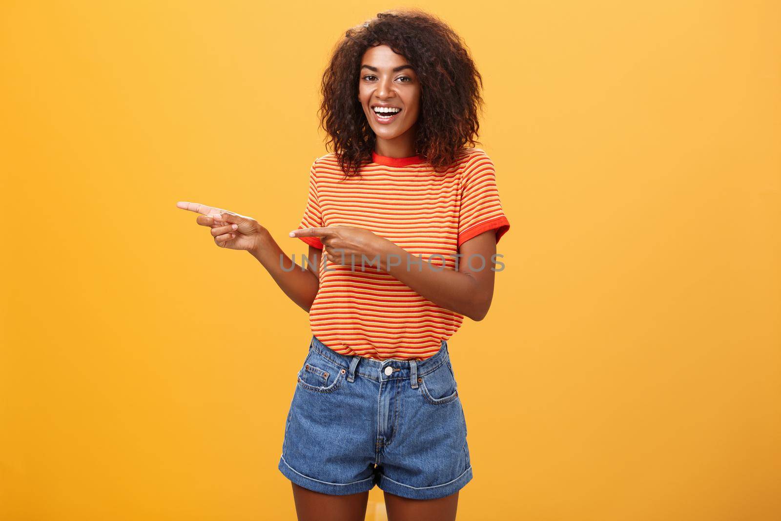 Ask him. Portrait of friendly and joyful good-looking stylish female shop assistant with curly hair and dark skin pointing left with both hands, smiling assured and entertained over orange background by Benzoix