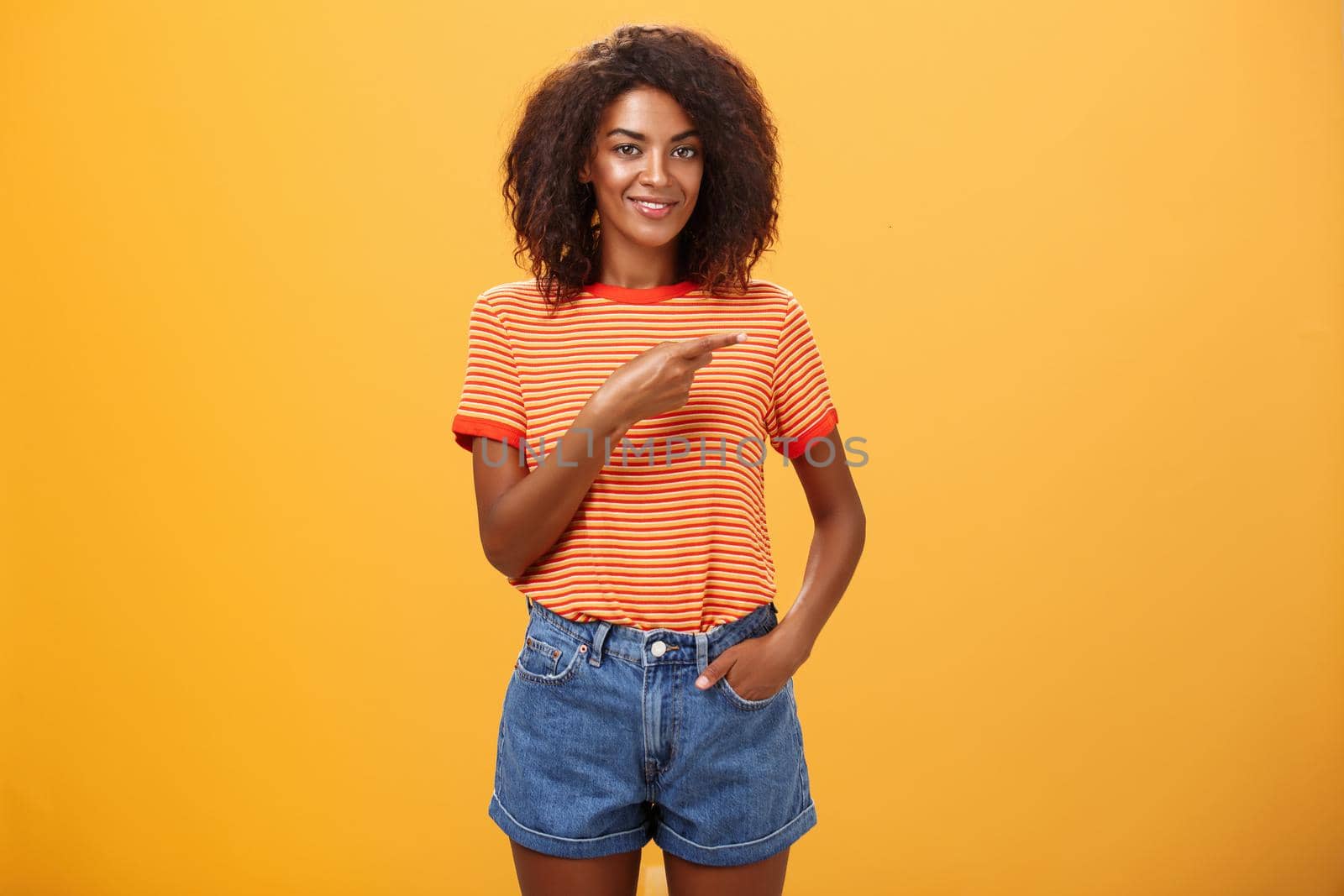 Showing perfect place for advertising. Charming carefree and confident young stylish african-american woman with afro hairstyle holding hand in pocket casually pointing right over orange wall.