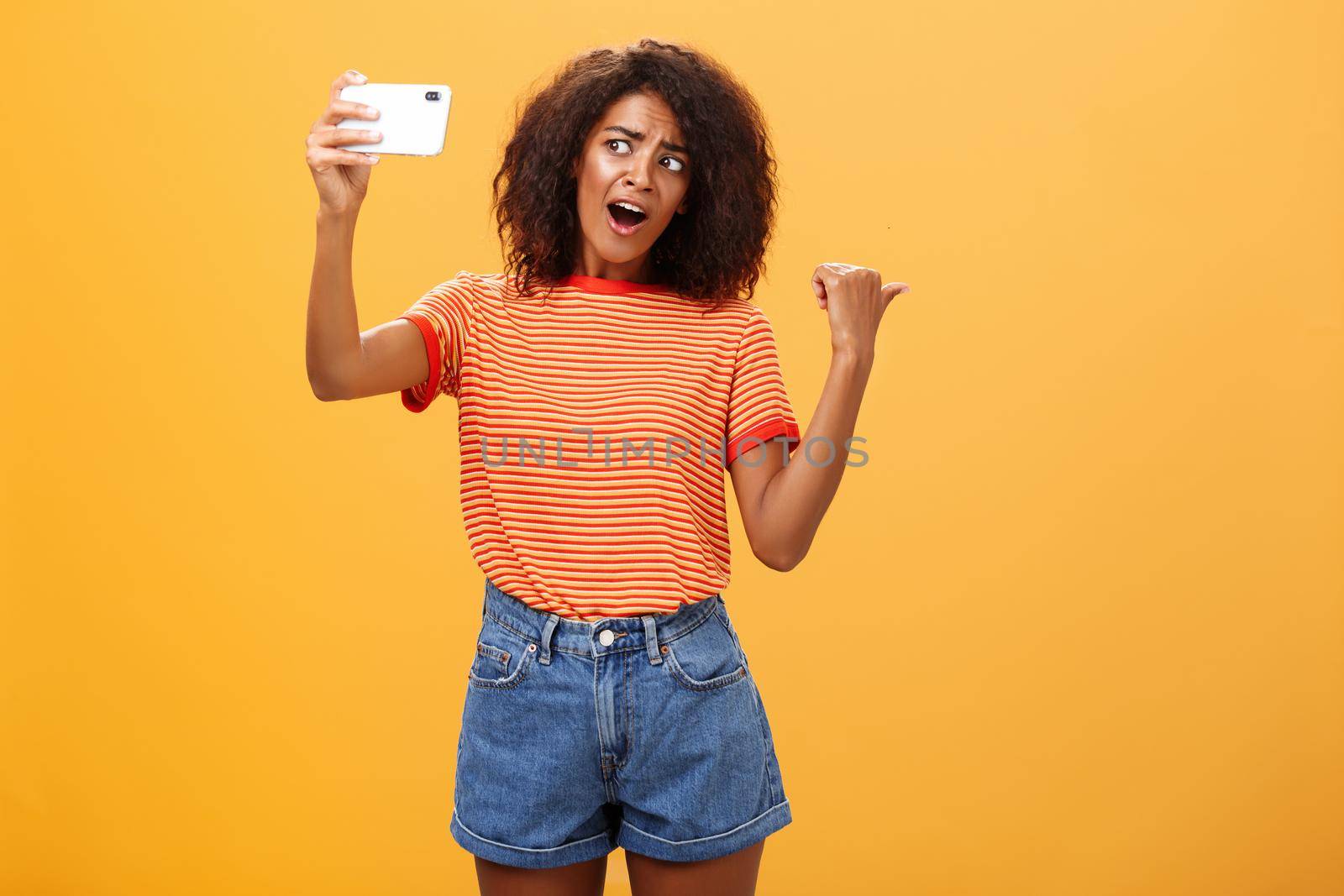 Woman recording video blog pointing at strange object behind her. Portrait of concerned and curious stylish famous internet star holding smartphone talking in cellphone camera over orange background. Technology concept