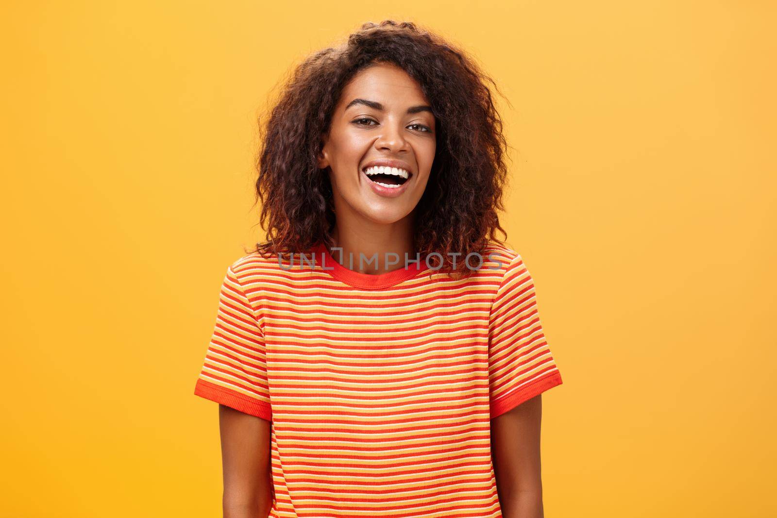 Waist-up shot of outgoing happy charming dark-skinned female with curly hairstyle laughing joyfully posing in striped trendy t-shirt over orange background enjoying nice casual conversation by Benzoix