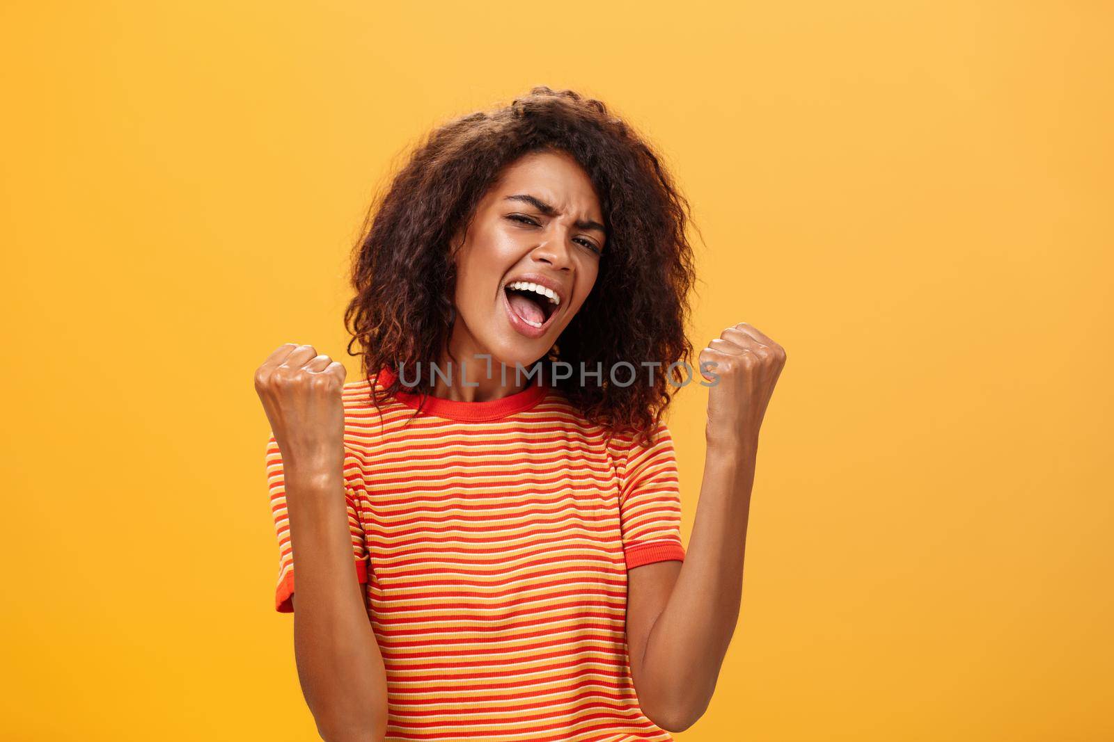 Cheerful delighted and enthusiastic african american woman with afro hairstyle clenching raised fists yelling yes from triumph and joy of success standing satisfied of victory over orange wall by Benzoix