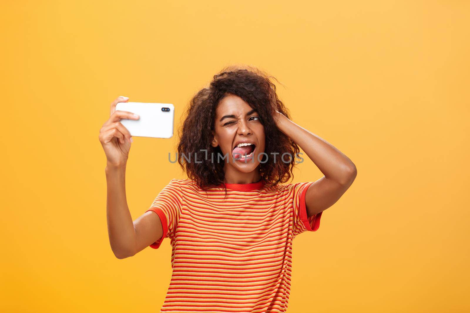 Waist-up shot of stylish confident african american woman with afro hairstyle in trendy t-shirt taking selfie on awesome new cellphone sticking out tongue playing with hair and winking at smartphone by Benzoix