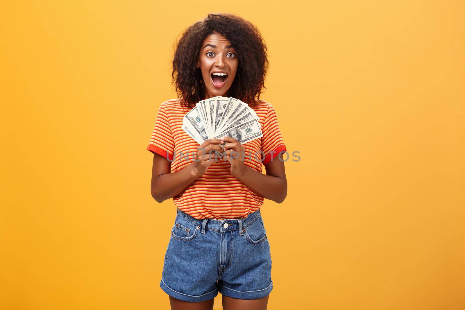 Woman cannot hide happiness winning lottery. Portrait of surprised and happy lucky african american young woman with curly hair holding bunch of money and yelling from delight receiving paycheck by Benzoix