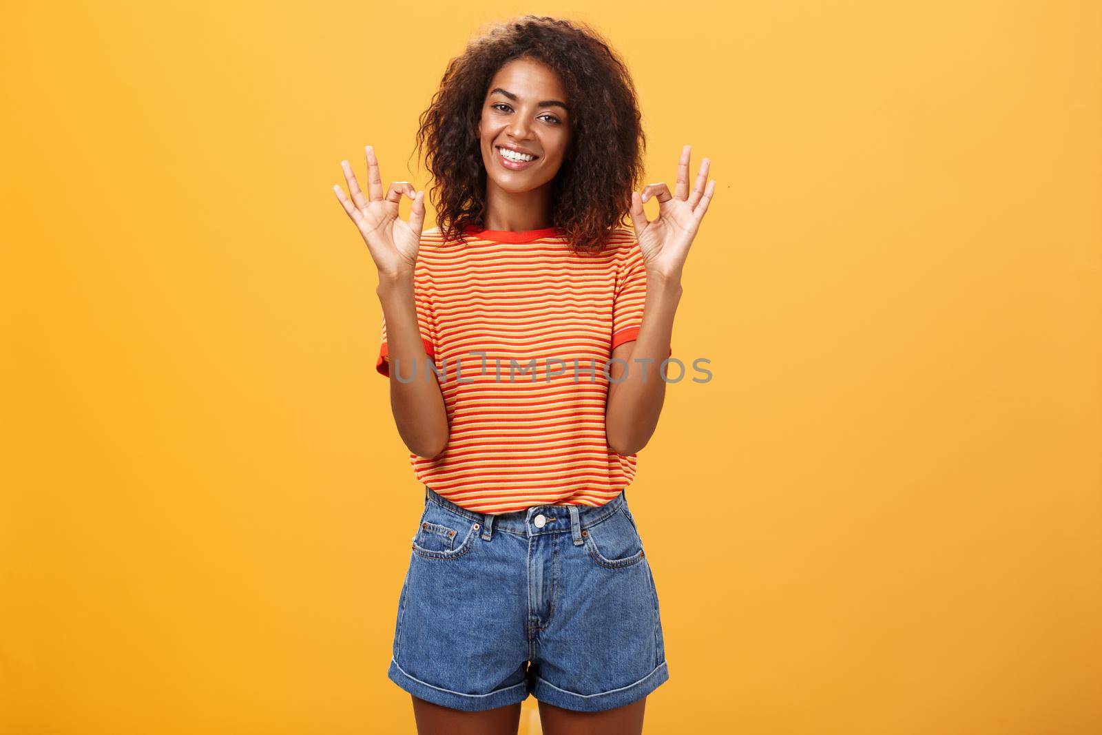 Chill attractive satisfied african american female customer picking new outfit being delighted and pleased with cool staff raising hands in okay or awesome gesture smiling happily over orange wall. Lifestyle.