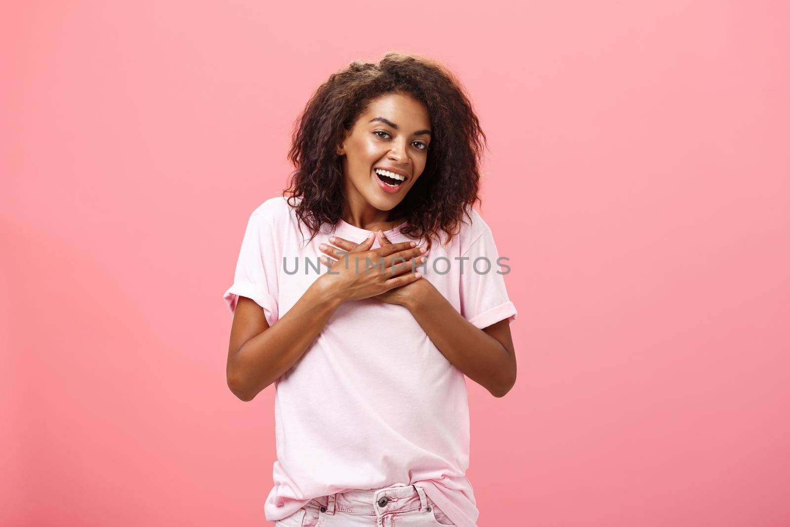 Portrait of charming delighted african american woman with curly haircut holding palms on heart pleased and grateful thanking friend for help smiling happily and thankful at camera over pink wall. Lifestyle.