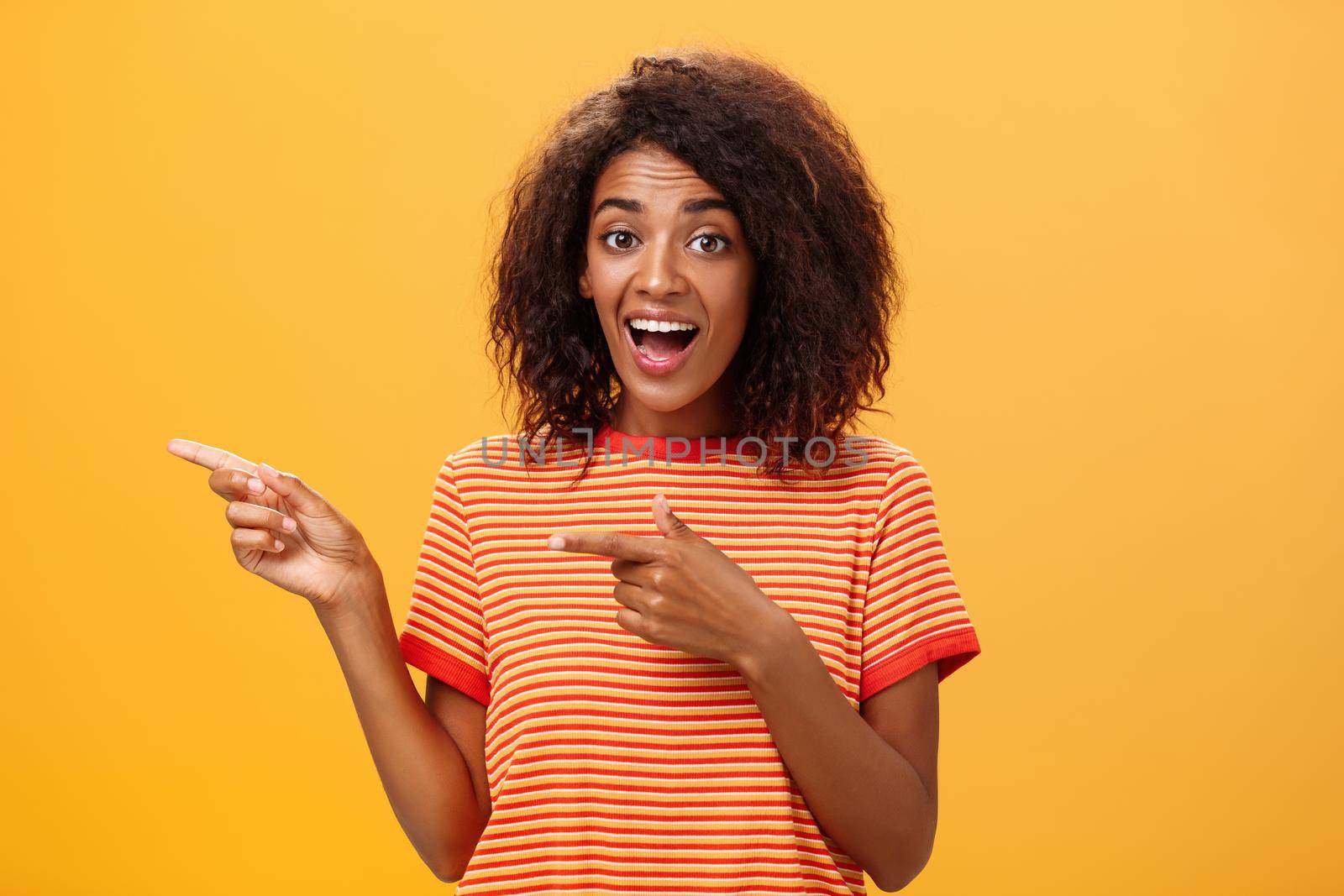 Portrait of amazed excited charismatic dark-skinned young pretty girl with afro hairstyle in trendy striped t-shirt pointing left delighted and fascinated posing against orange background. Lifestyle.