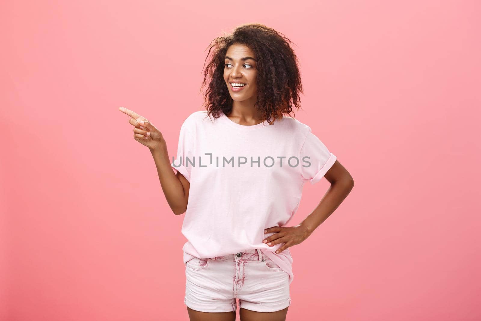 Girl checking out nice body of guy passing by. Charming confident dark-skinned female with curly hairstyle holding hand on hip looking and pointing left discussing cool copy space over pink wall by Benzoix