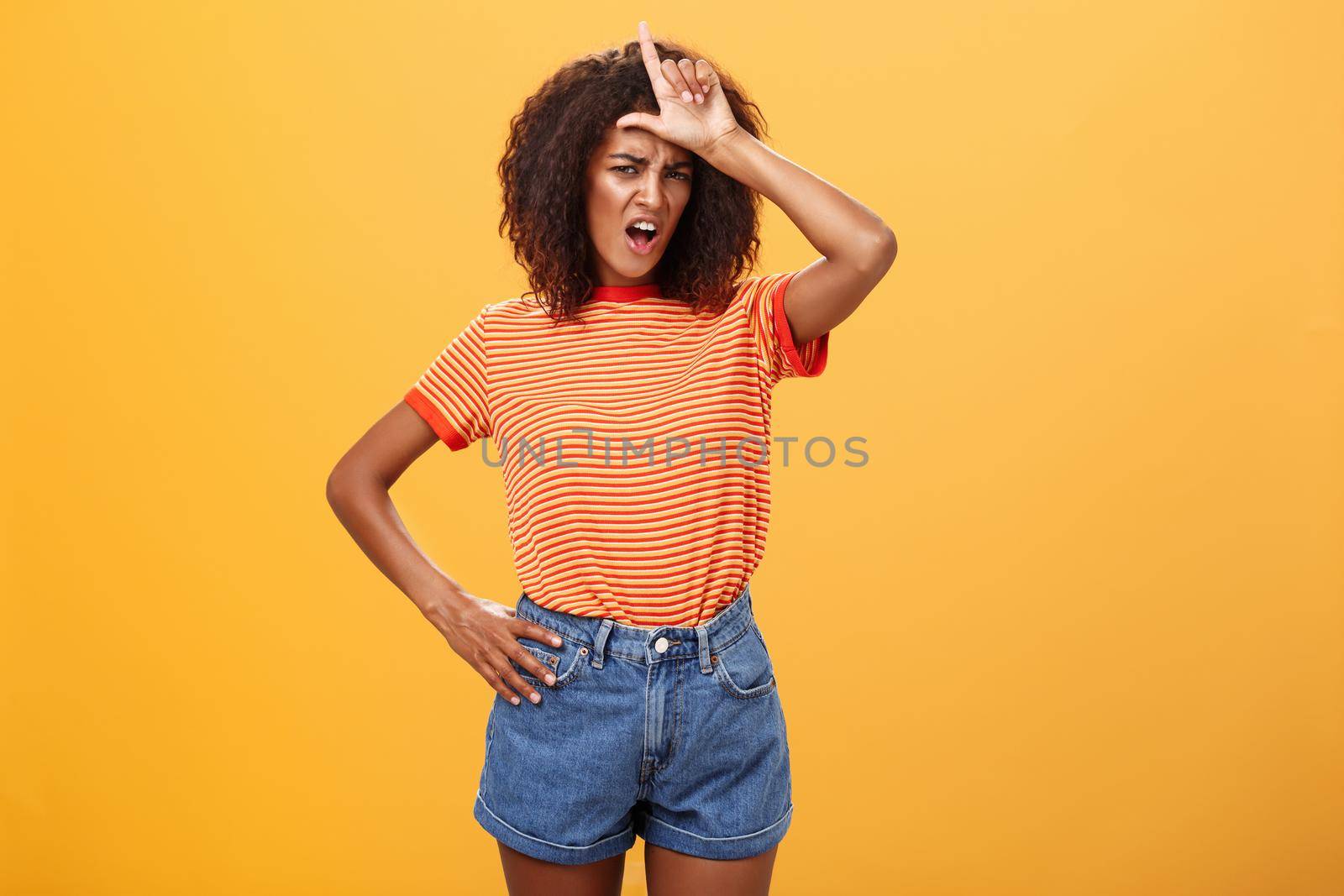 Arrogant and confident woman not talk with losers. Mocking self-assured stylish african american female with curly hair in trendy clothes frowning scolding nerd, showing letter l on forehead. Copy space