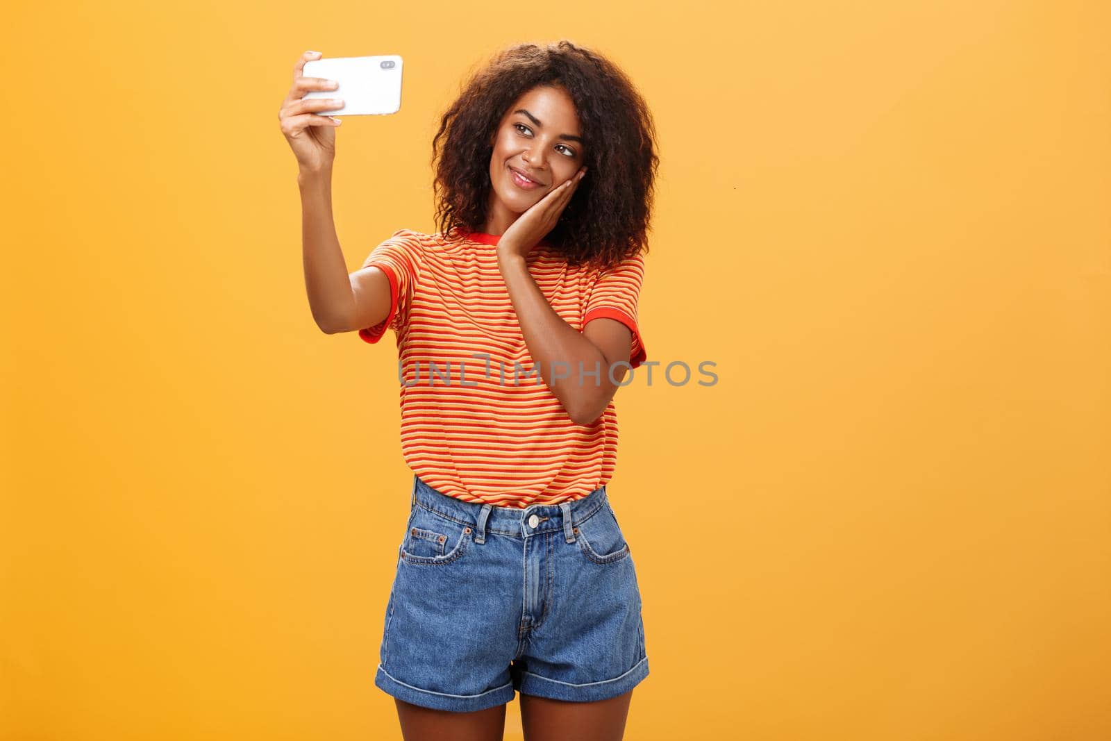 Woman made make-up all day to post new photo online taking selfie on brand new smartphone making cute face leaning head on palm smiling gently at device screen posing over orange background. Technology and lifestyle concept