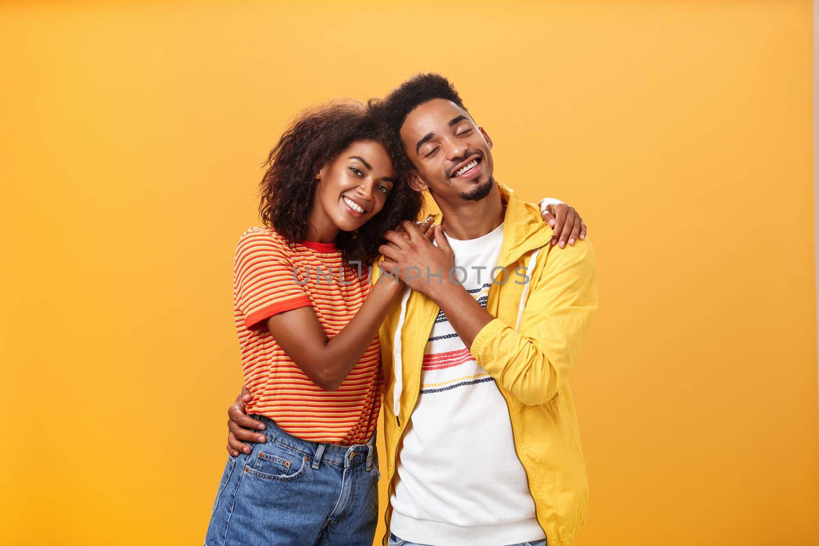 They love each other. Portrait of two charming african american man and woman in relationship hugging with heartwarming smile touching hands smiling gently posing against orange background.