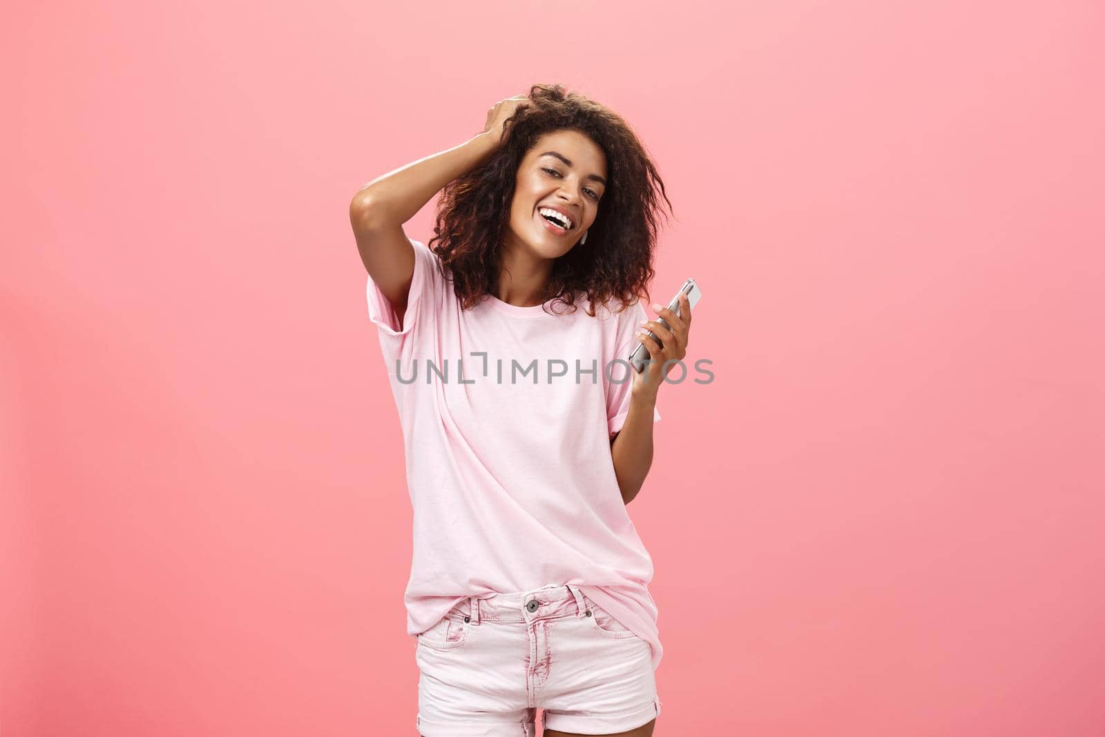 Portrait of carefree stylish modern dark-skinned young girl using smartphone hanging around joyfully touching hair and gazing with broad smile at camera holding cellphone posing against pink wall. Technology concept