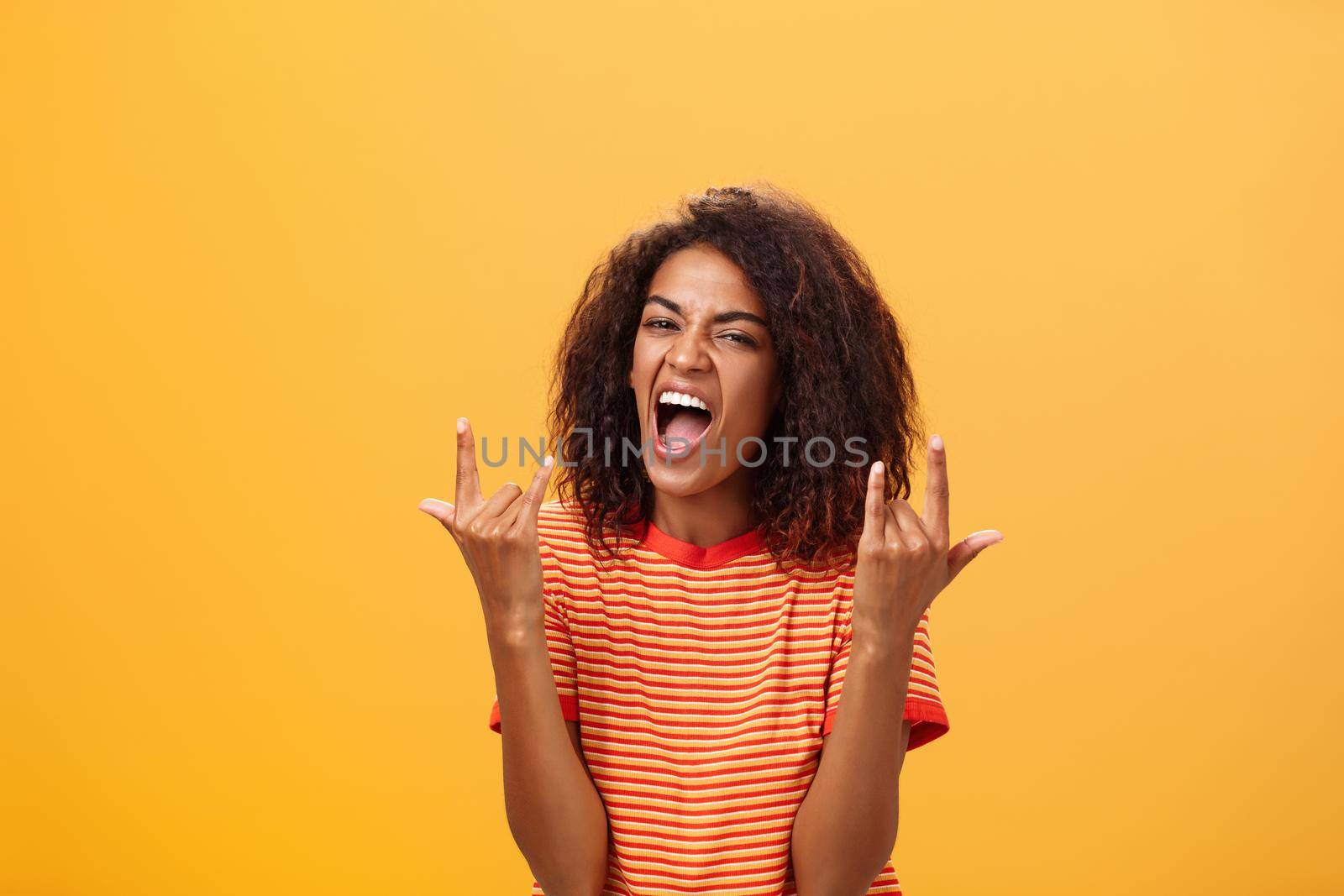 Waist-up shot of amazed happy stylish african american woman feeling awesome rocking on party yelling from joy and satisfaction showing rock n roll gesture posing over orange background. Copy space