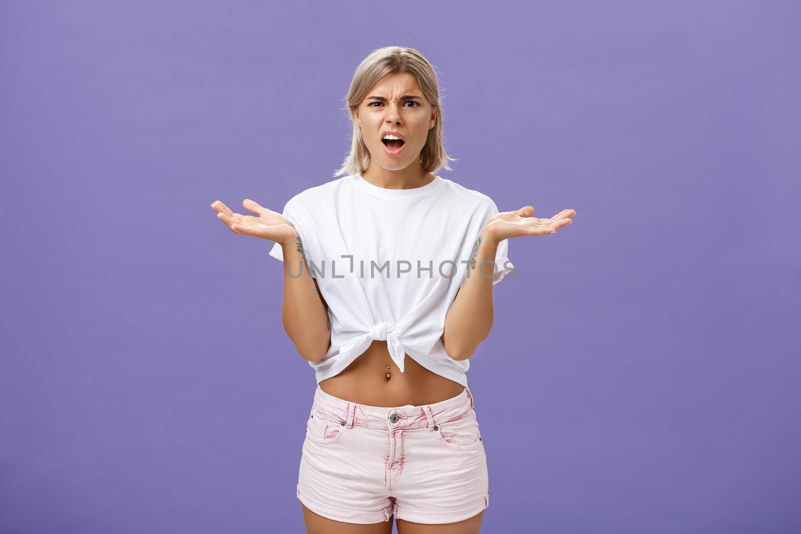 Perplexed and disappointment good-looking blonde female student in white t-shirt and pink shorts frowning shrugging with spread hands near shoulders saying what the hell over purple wall by Benzoix