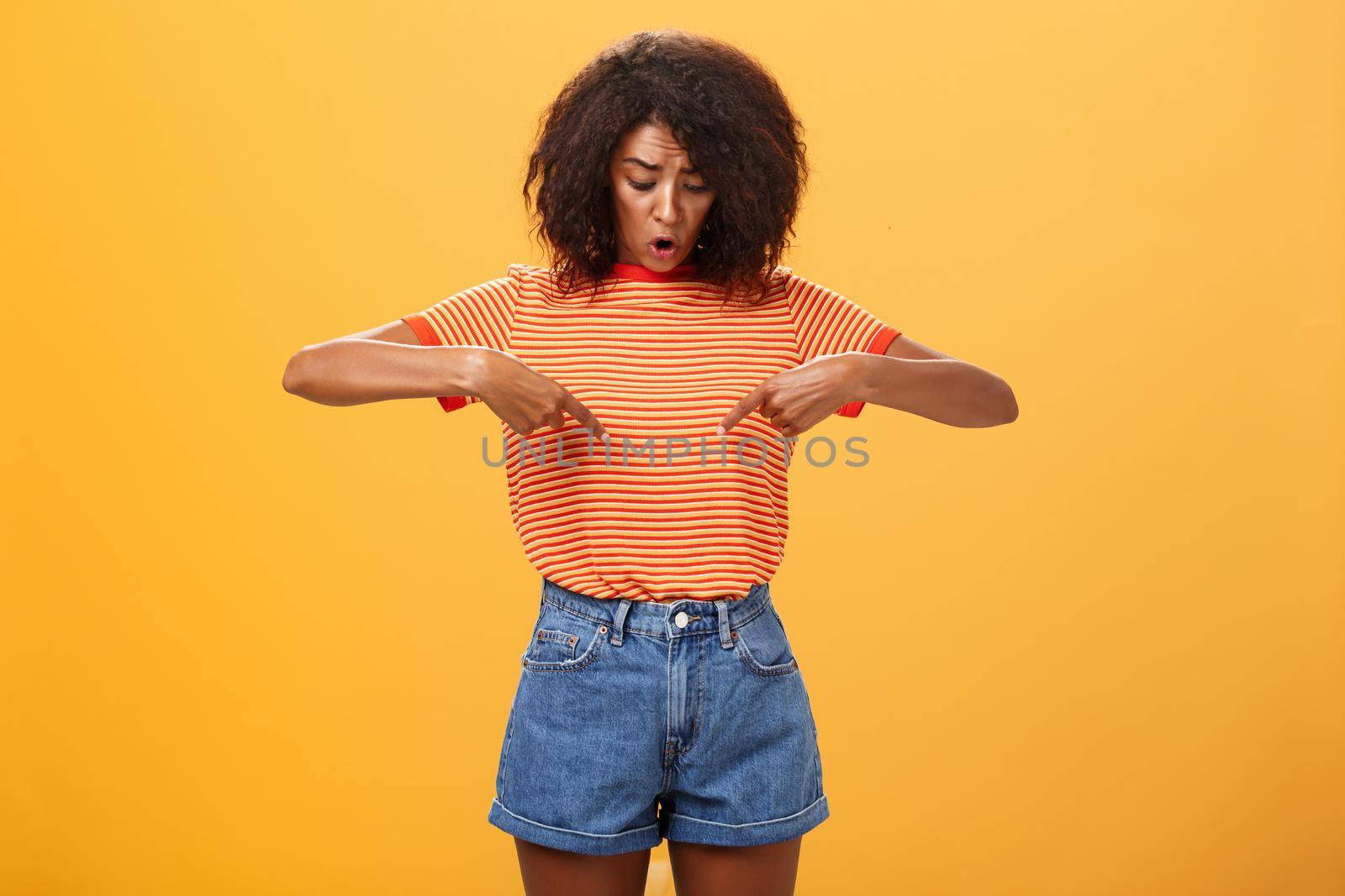 Portrait of worried questioned and surprised african american female with curly hairstyle looking and pointing at stomach or belly feeling discomfort and problems with health over orange background. Lifestyle.