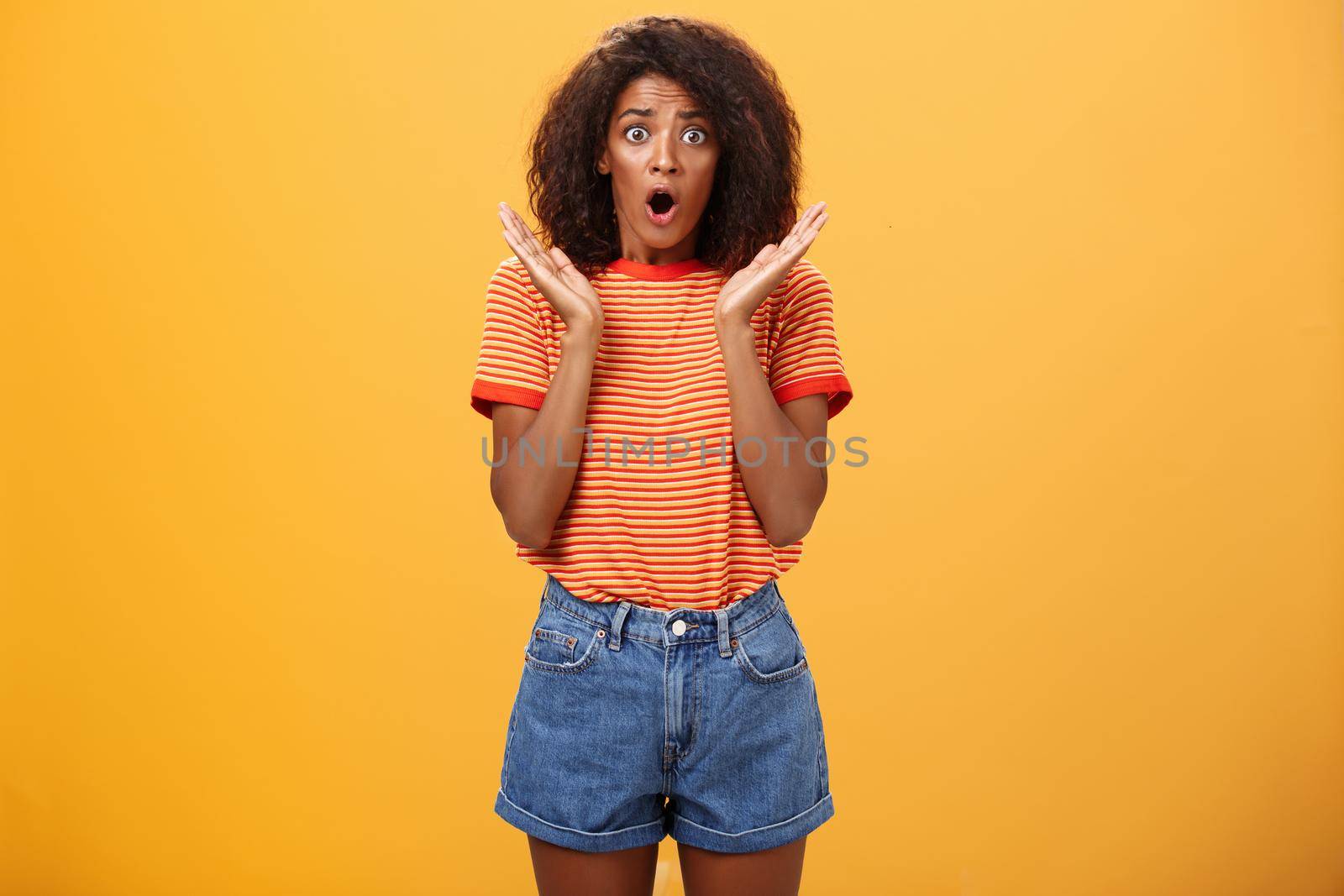 Oh no what happened. Worried african american sister with curly hair in trendy striped t-shirt and denim shorts gasping dropping jaw, looking concerned expressing empathy and shock over orange wall.