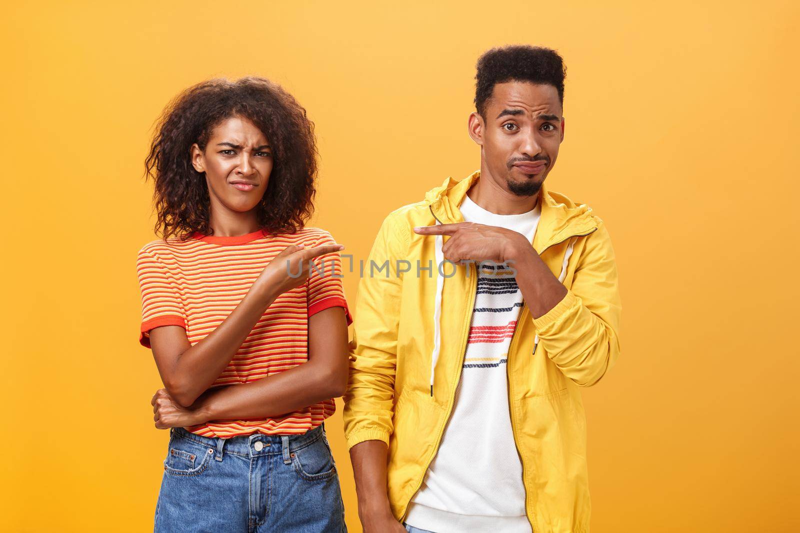 Friends getting shocked someone thought they are dating. Displeased mocking african american female frowning and grimacing from aversion while pointing at guy who indicating at girl over orange wall by Benzoix