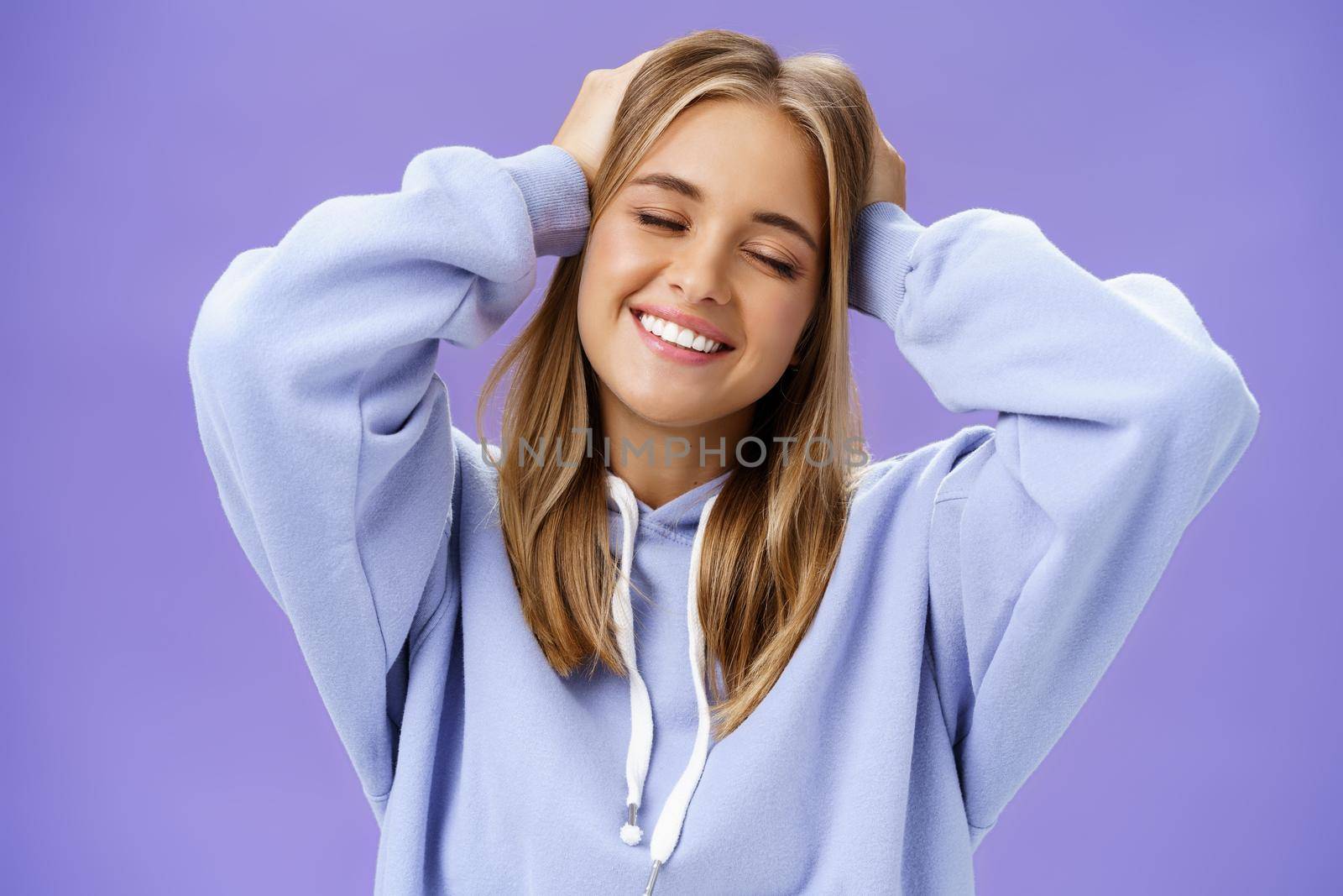 Tender relaxed charming woman with blond hair touching head joyfully smiling broadly with closed eyes daydreaming posing in trendy hoodie against purple wall finally getting rid of acne and pimples by Benzoix