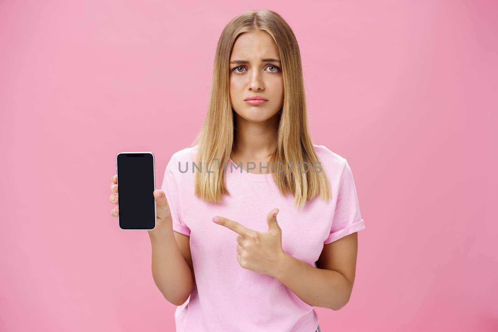 Gloomy and sad cute young female showing friend negative feedback about her project in internet pointing at smartphone screen standing concerned and upset against pink background by Benzoix