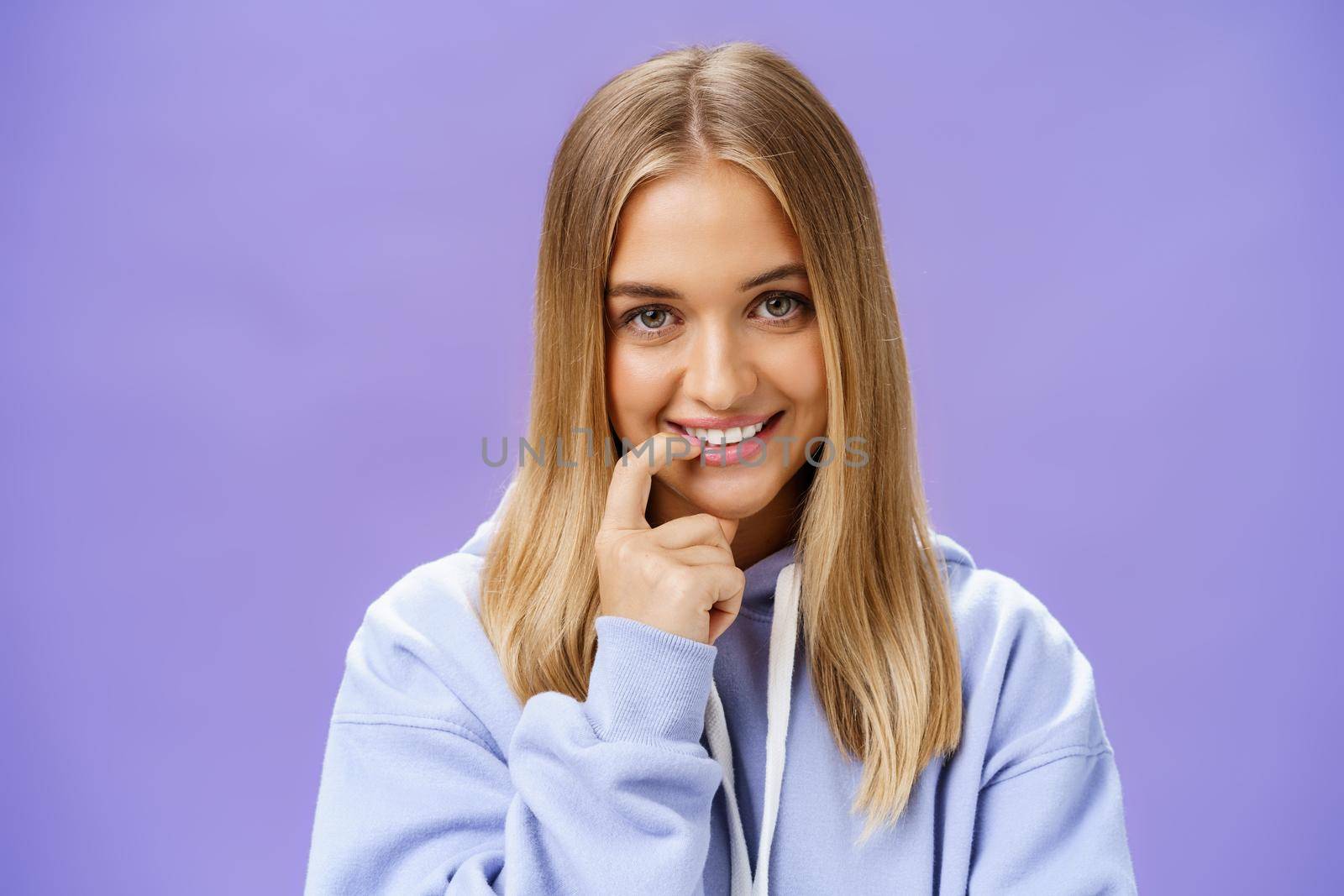 Close-up shot of feminine and tender young girl with fair hair and tanned skin biting finger flirty and shy smiling cute at camera standing in trendy over-sized hoodie, flirting over purple background. Emotions concept