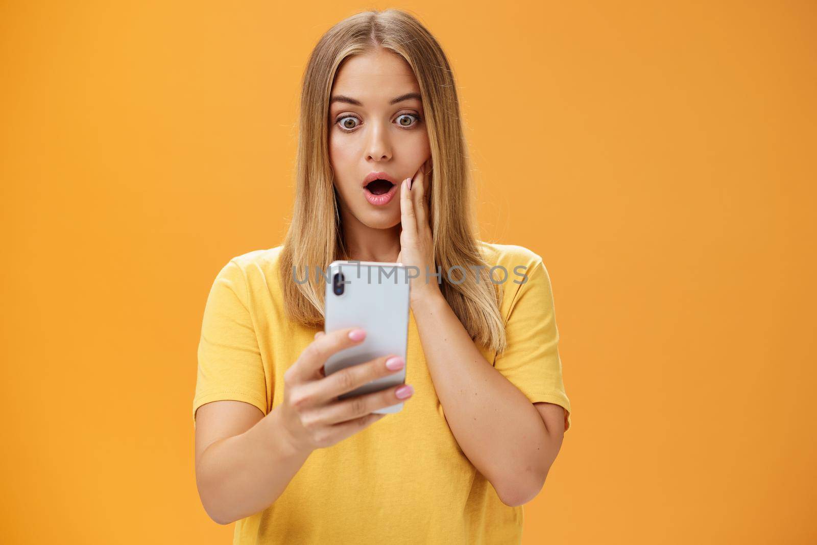 Waist-up shot of shocked and stunned emotive young woman reacting to shocking terrible news reading online from internet gasping opening mouth nervously touchin cheek staring at smartphone screen. Technology concept