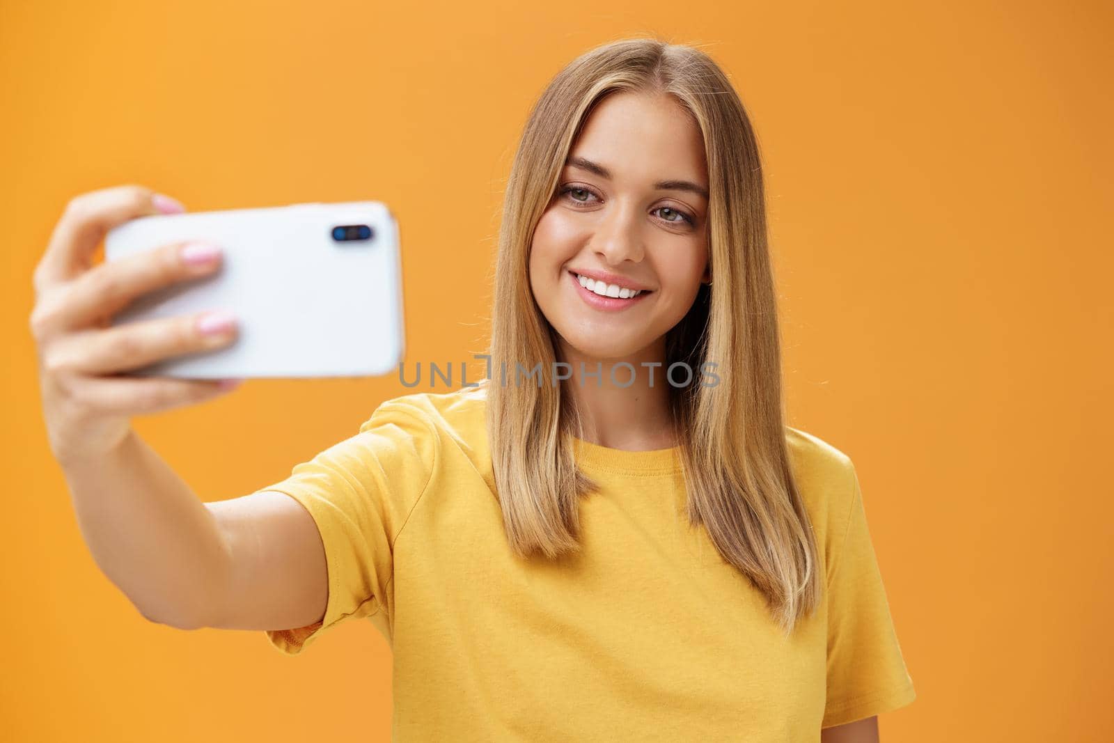 Waist-up shto of outgoing and charismatic pretty confident woman with no makeup and tanned skin holding smartphone pulled on distance smiling broadly at cellphone screen while taking selfie. Technology concept