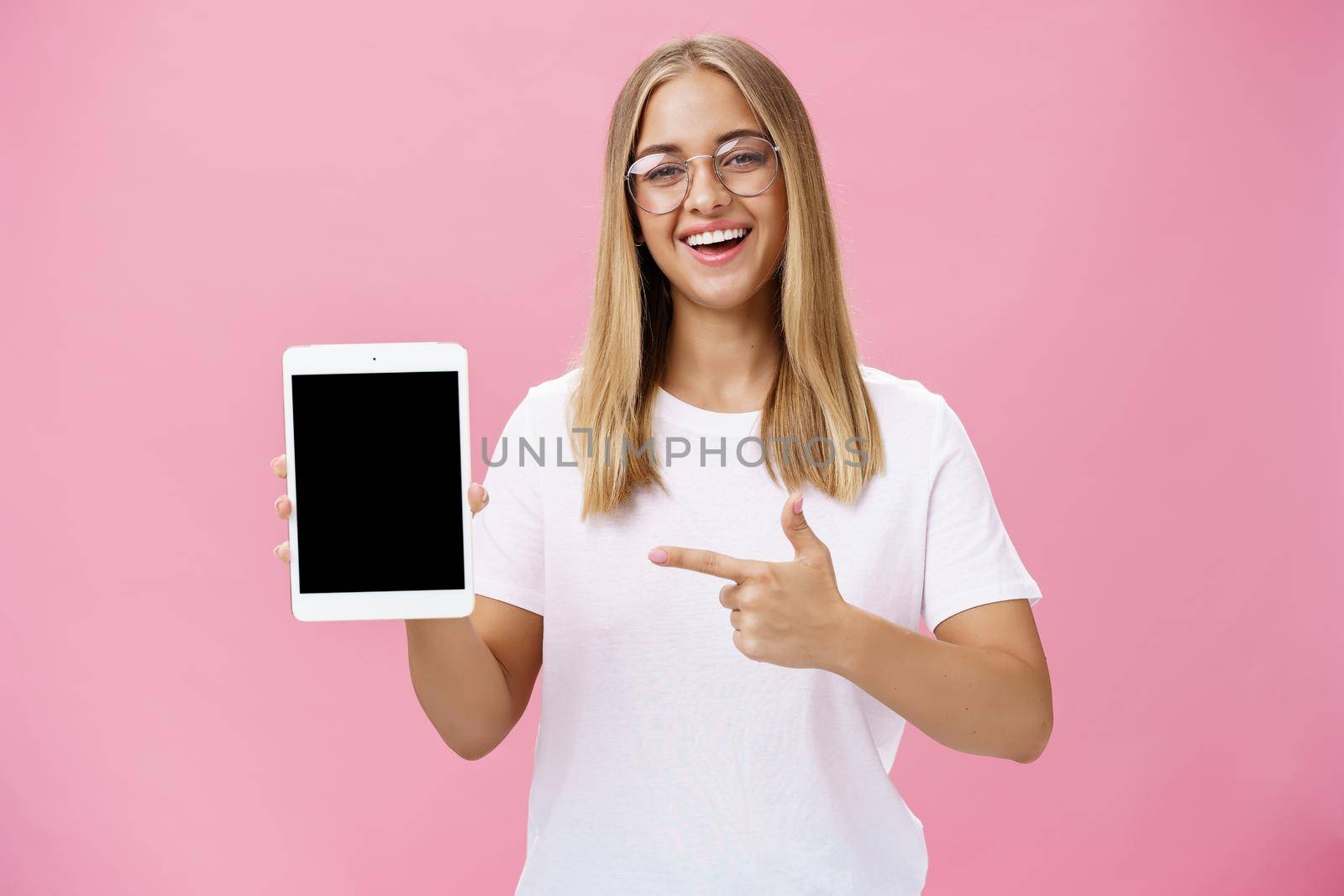 Female freelance programmer proudly showing her app for digital tablet holding gadget pointing at device screen smiling broadly with delighted expression wearing glasses, posing over pink wall. Technology and advertising concept