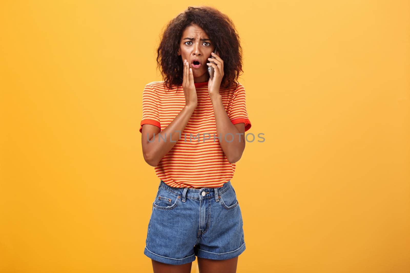 Woman receiving disturbing call feeling emapthy and sorry for poor friend getting in trouble holding cellphone near ear touching cheek concerned frowning standing sad, worried over orange background.