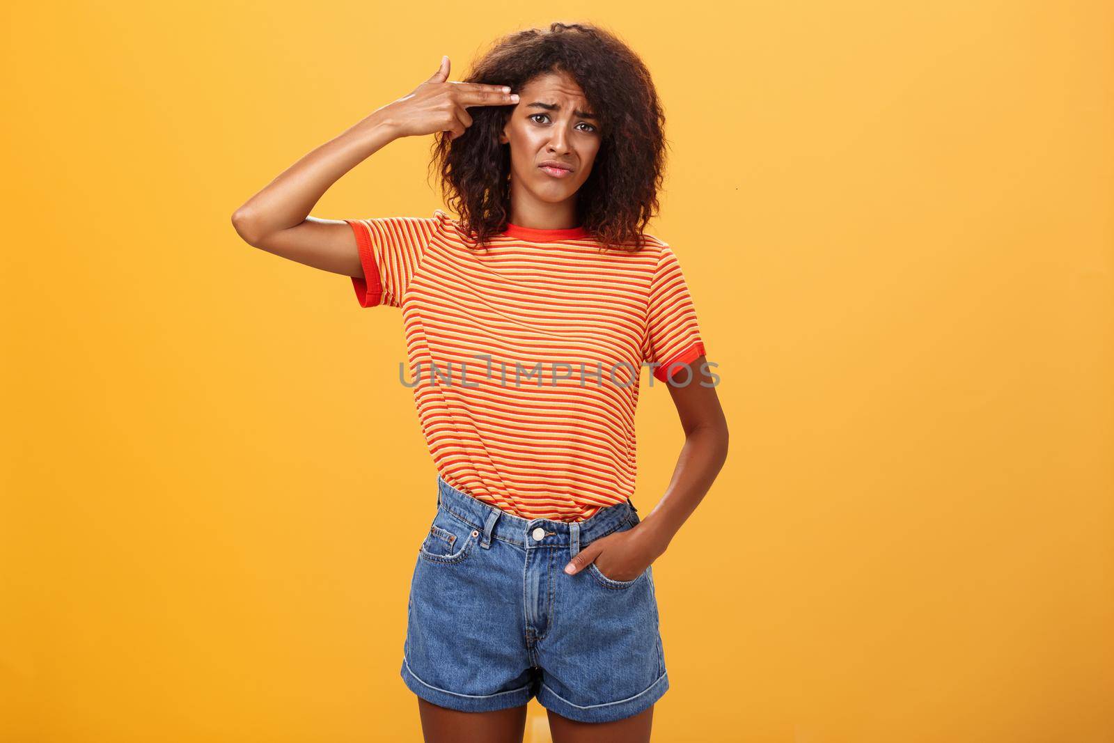 Portrait of displeased gloomy and sad annoyed dark-skinned female with curly hairstyle showing gun with hand near temple frowning wanting to kill herself from exhaust and boredom over orange wall. Lifestyle.