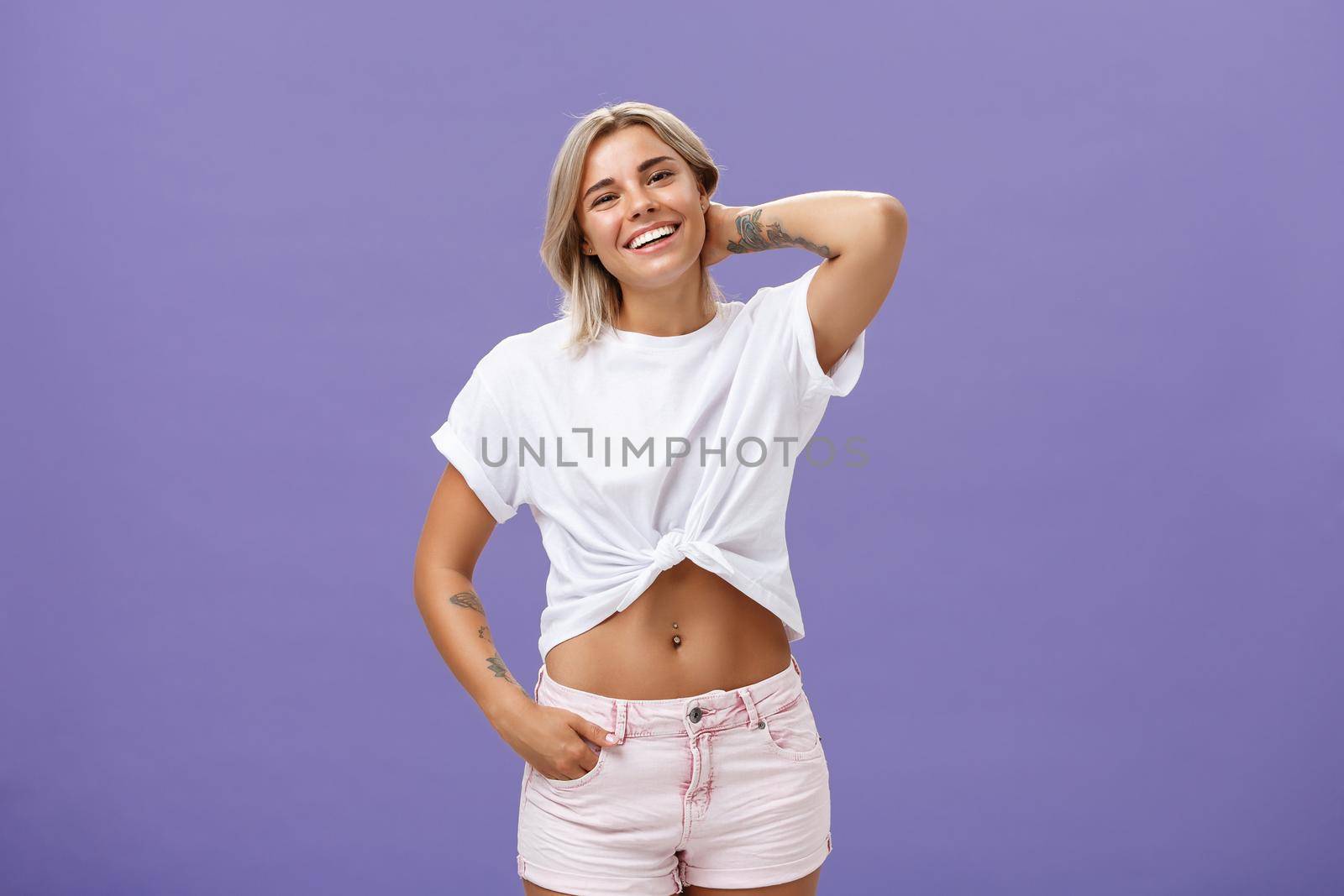 Lifestyle. Indoor shot of carefree relaxed and joyful attractive athletic blonde woman in trendy summer outfit touching back of neck and smiling joyfully as if being in awkward situation over purple wall.