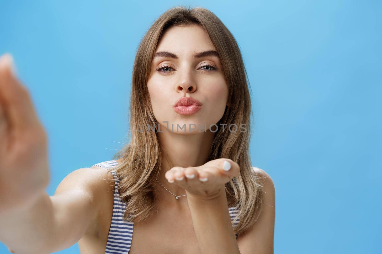 Close-up shot of self-assured glamorous and attractive stylish woman holding hand near folded lips to send kiss while grabbing camera and taking selfie standing sensual and romantic over blue wall. Lifestyle, network and people concept