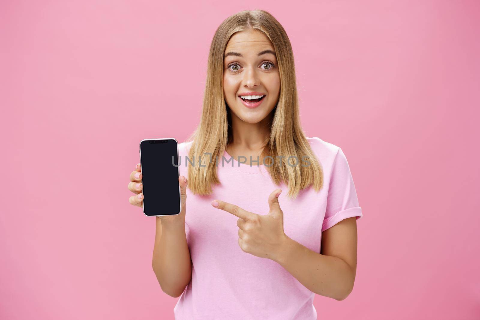 Cheerful attractive and pleasant woman promoting cool app or smartphone holding cellphone and pointing at device screen smiling amused and impressed standing over pink background by Benzoix