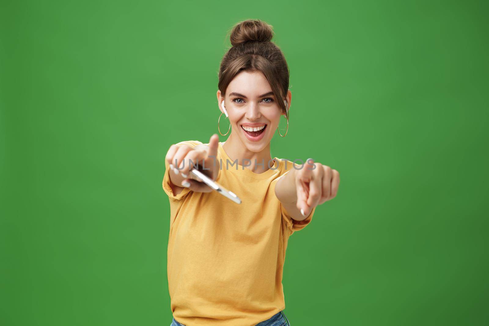 Energized charismatic attractive young woman with combed hair and cute diastema holding smartphone dancing joyfully listening music in wireless earphones pointing and smiling at camera over green wall by Benzoix