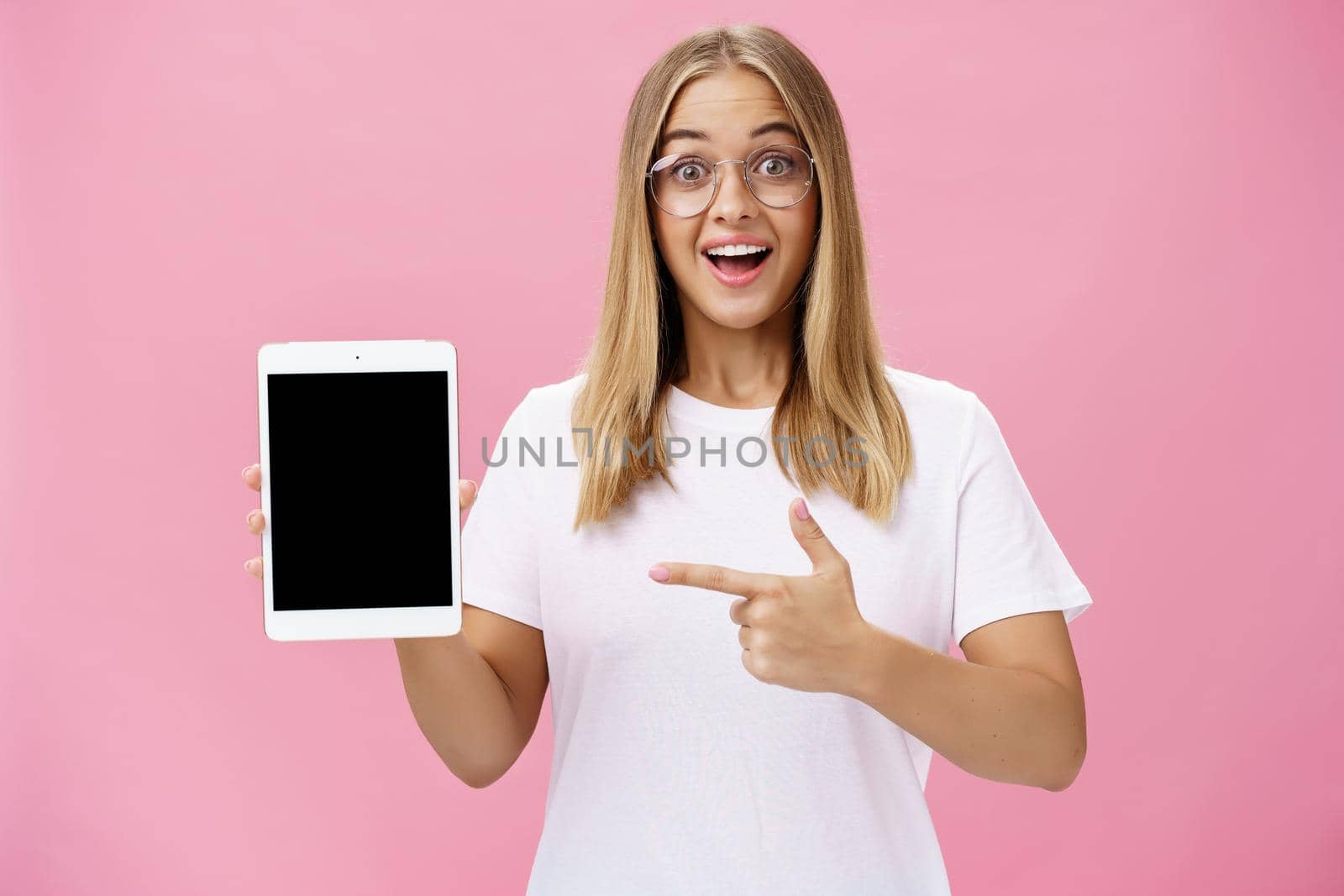 Girl suggests buy digital tablet for university and forget paper books. Excited happy and delighted young female student in white t-shirt and glasses pointing at device screen recommending gadget. Technology, emotions, advertising and lifestyle concept