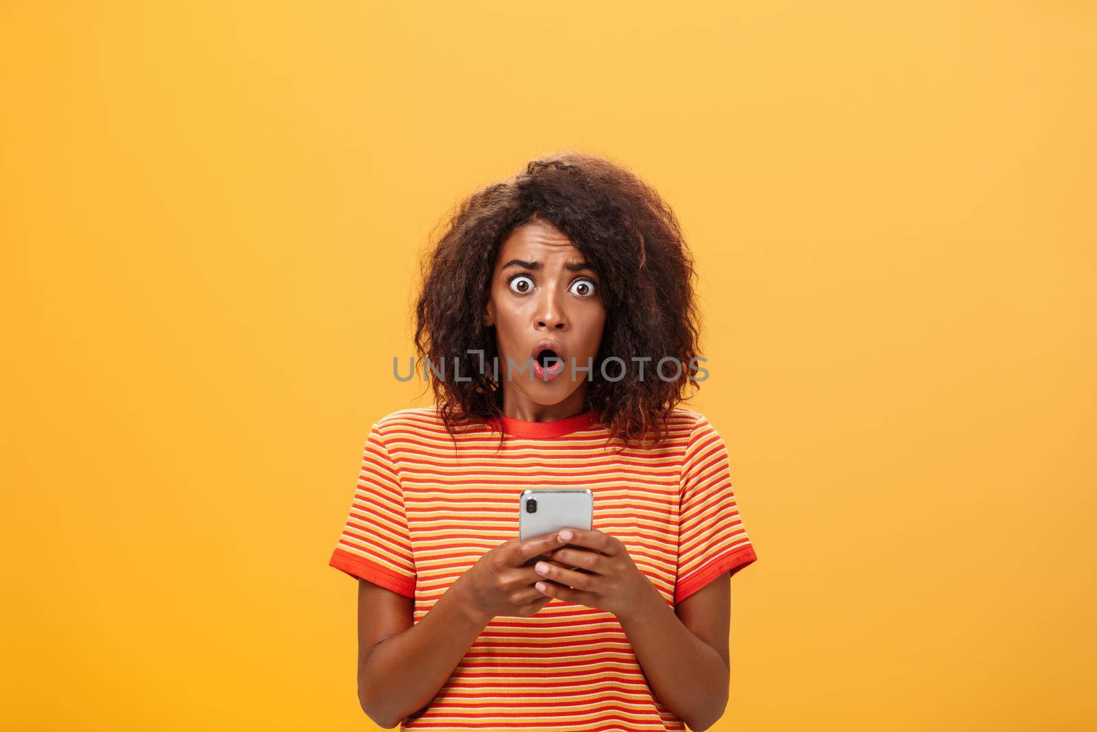 Portrait of shocked and concerned stunned dark-skinned woman with afro hairstyle popping eyes from concern and panic opening mouth holding smartphone revealing terrible information.