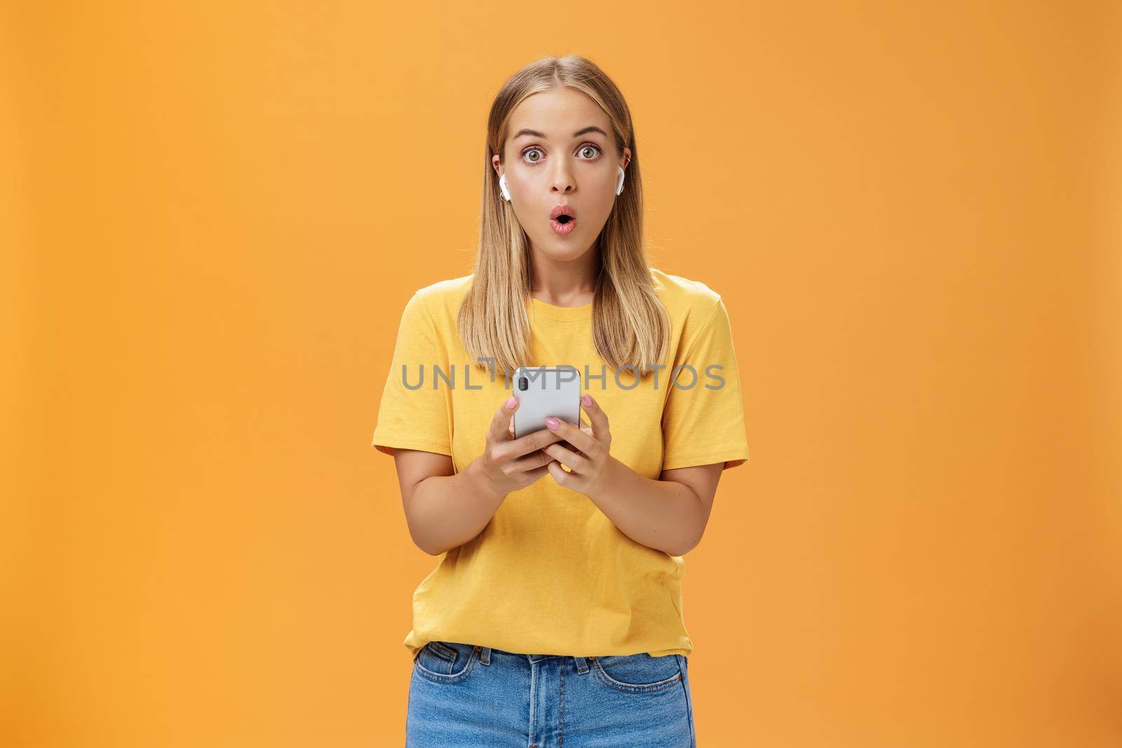 Woman surprised how awesome sounds new wireless earbuds holding smartphone wearing earphones folding lips in wow and amazement listening music impressed by good product over orange background. Copy space