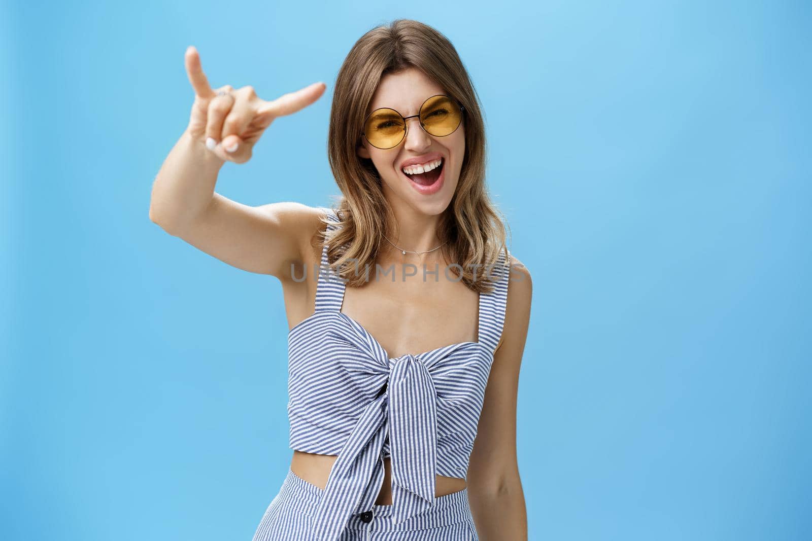 Waist-up shot of confident and daring charming young woman in matching trendy clothes yelling yeah showing rock-n-roll gesture and gazing at camera wearing cool sunglasses enjoying party or festival by Benzoix