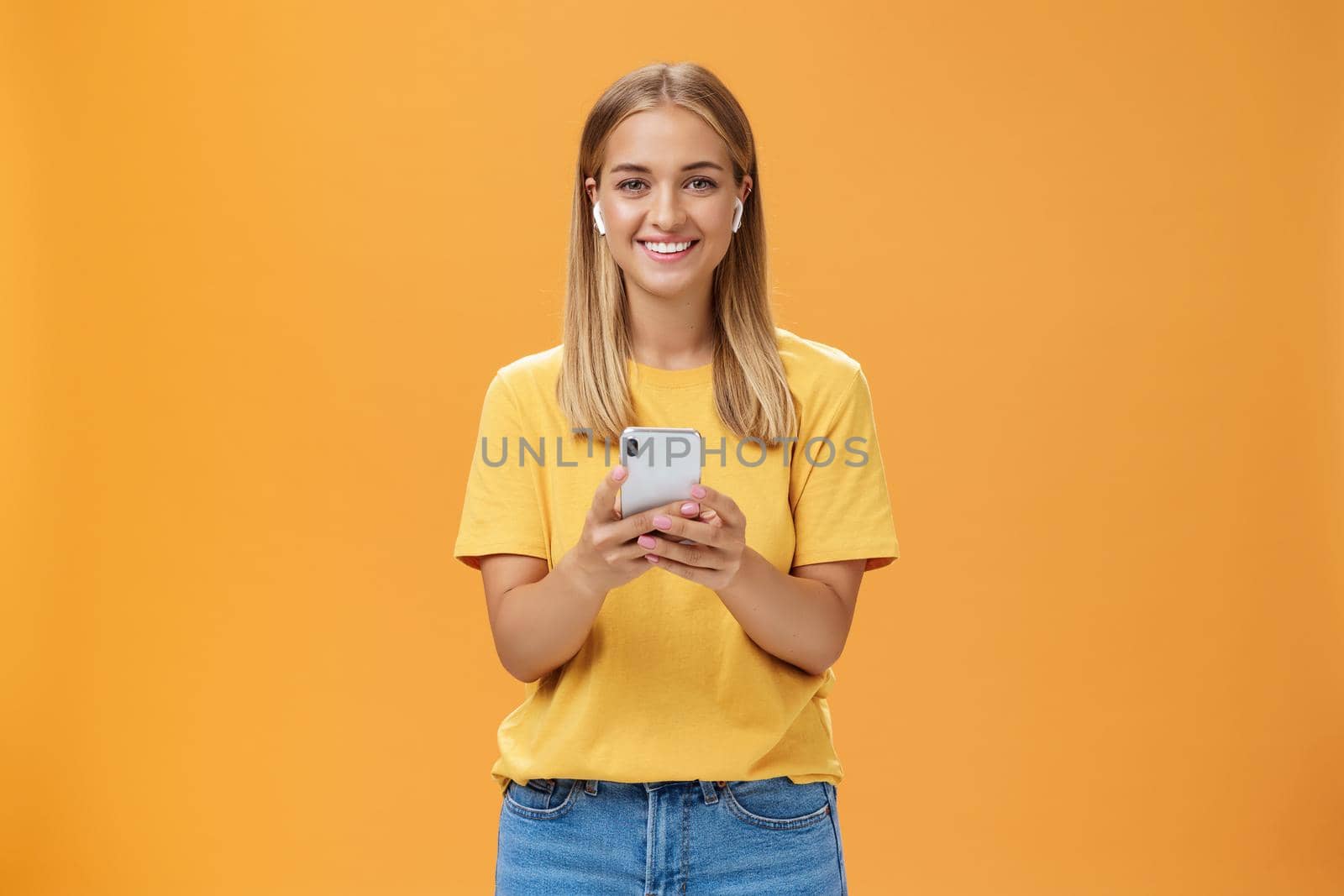 Portrait of friendly charismatic and outgoing good-looking young female in yellow t-shirt holding smartphone wearing wireless earphones enjoying listening music on way to fitness gym by Benzoix