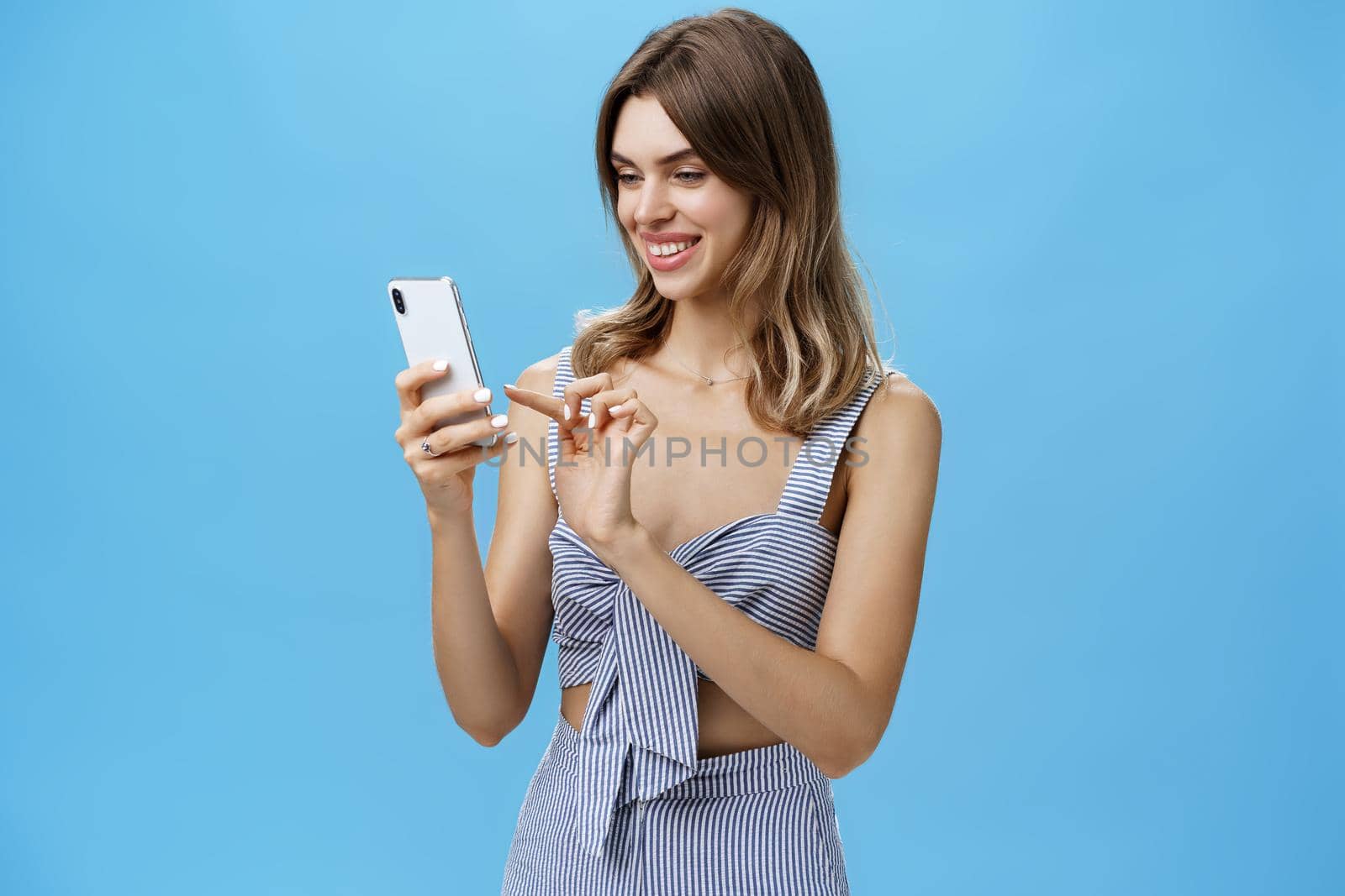 Portrait of stylish sociable good-looking female entrepreneur making appointment via smartphone holding cellphone typing message gazing at device screen with pleased carefree smile over blue wall by Benzoix