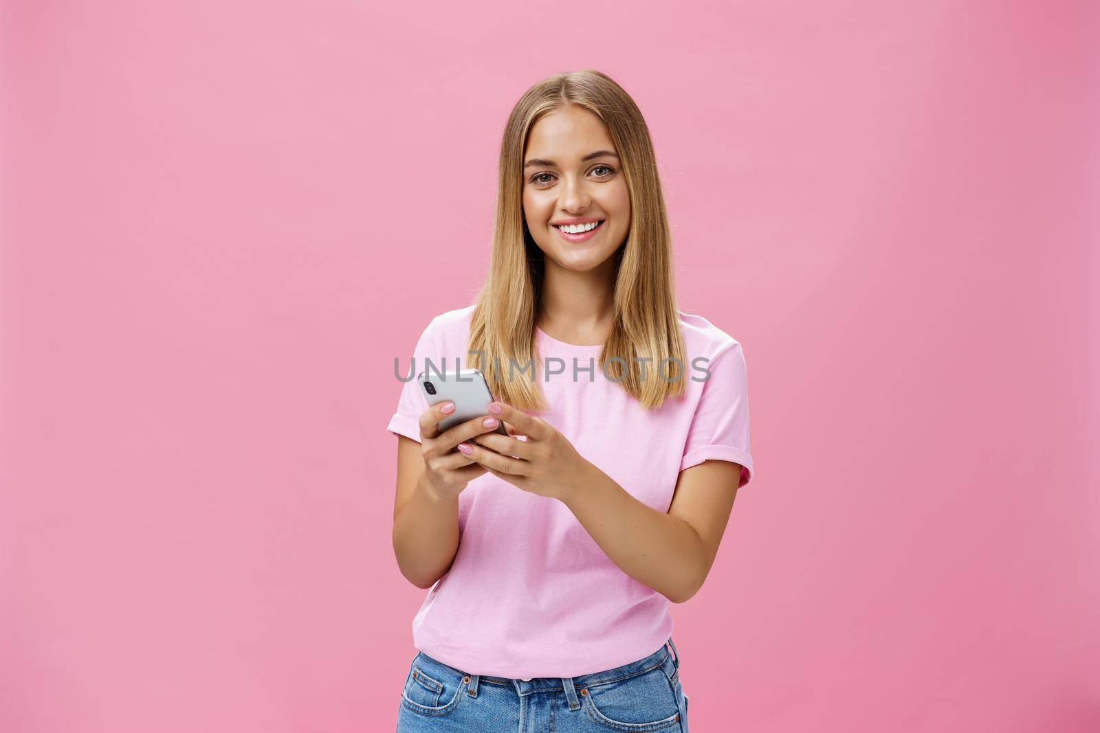 Woman calling taxy via smartphone asking friend address smiling cheerfully at camera holding phone with both hands over chest getting in touch with clients via messages standing over pink wall by Benzoix