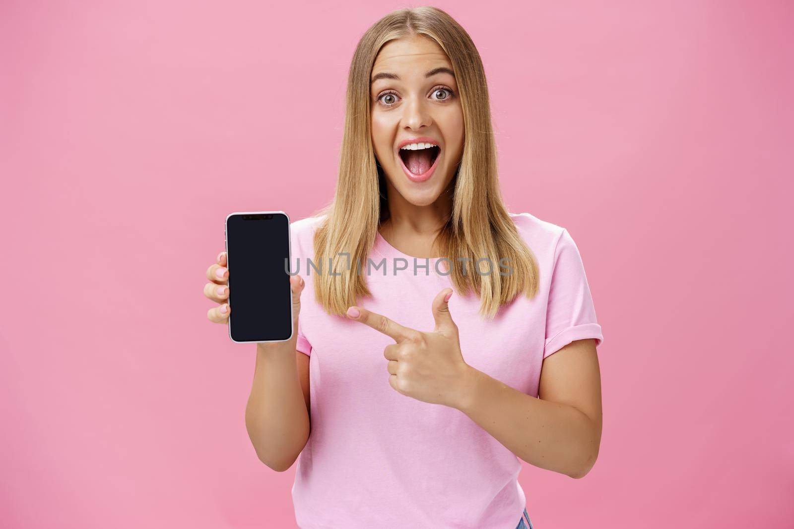 Portrait of excited and amused charming pleased female client using new smartphone advicing friends to buy awesome gadget holding cellphone and pointing at device screen with broad amused smile by Benzoix